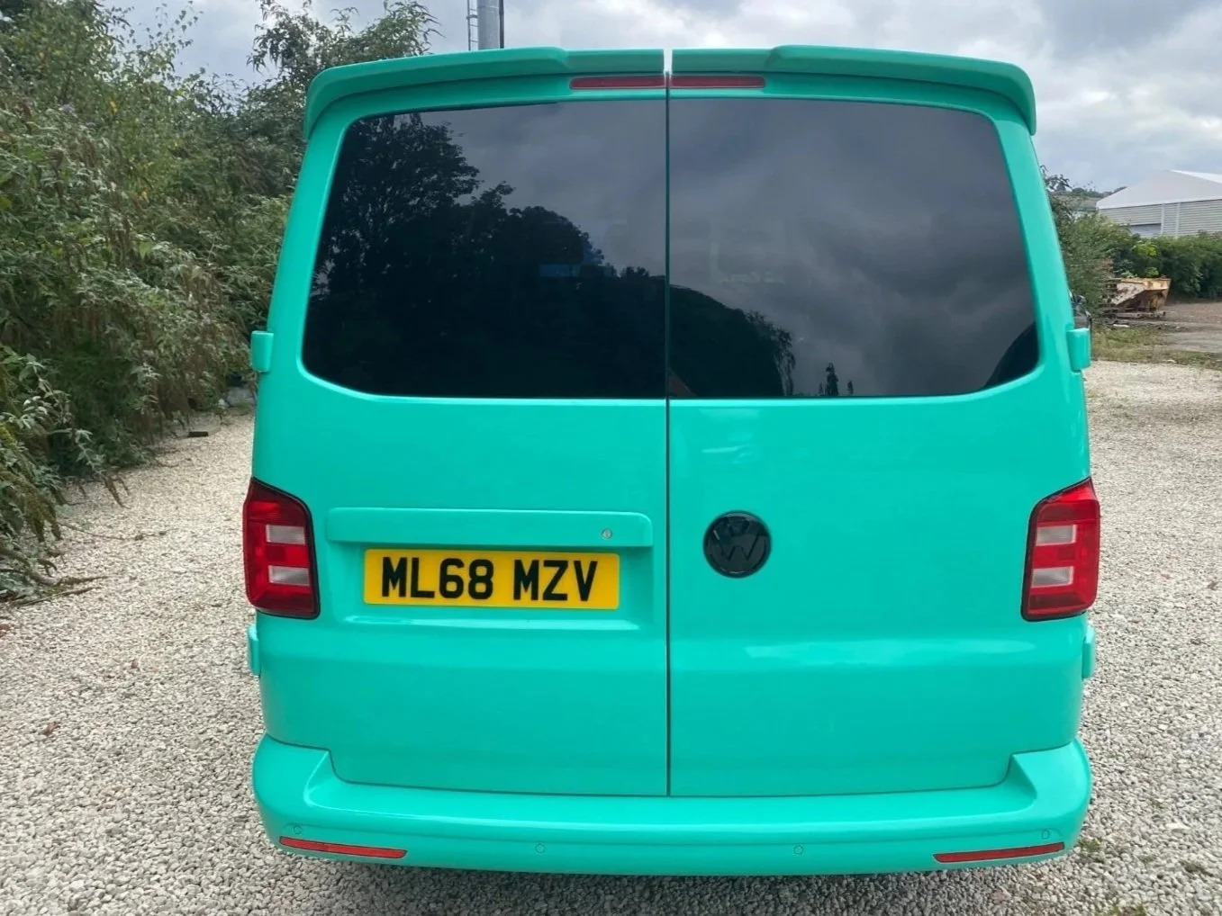 Rear view of a teal-colored Volkswagen van with a yellow license plate reading 'ML68 MZV', parked on gravel with greenery on the left and some buildings in the background.