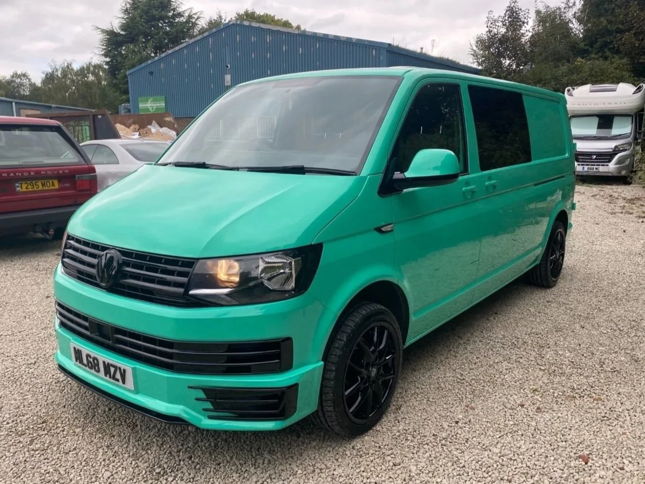 A mint green Volkswagen commercial van parked on a gravel lot, with a black front grille and black alloy wheels.