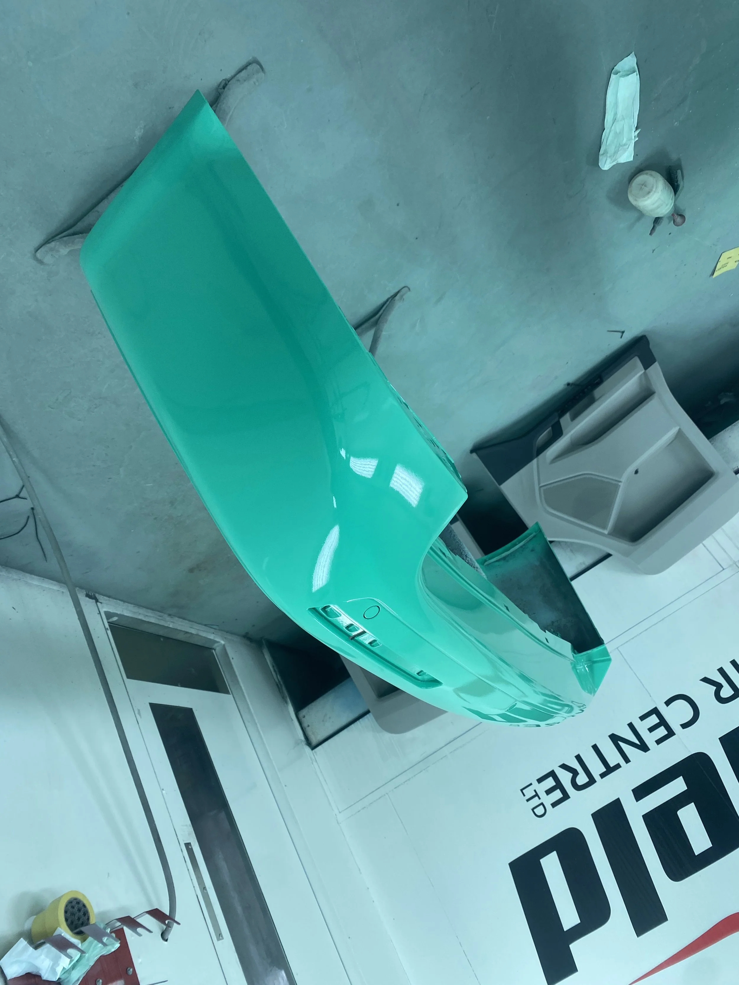 A teal-colored car hood placed on a table in a workshop or garage. Tools, paper towels, and containers are visible nearby.