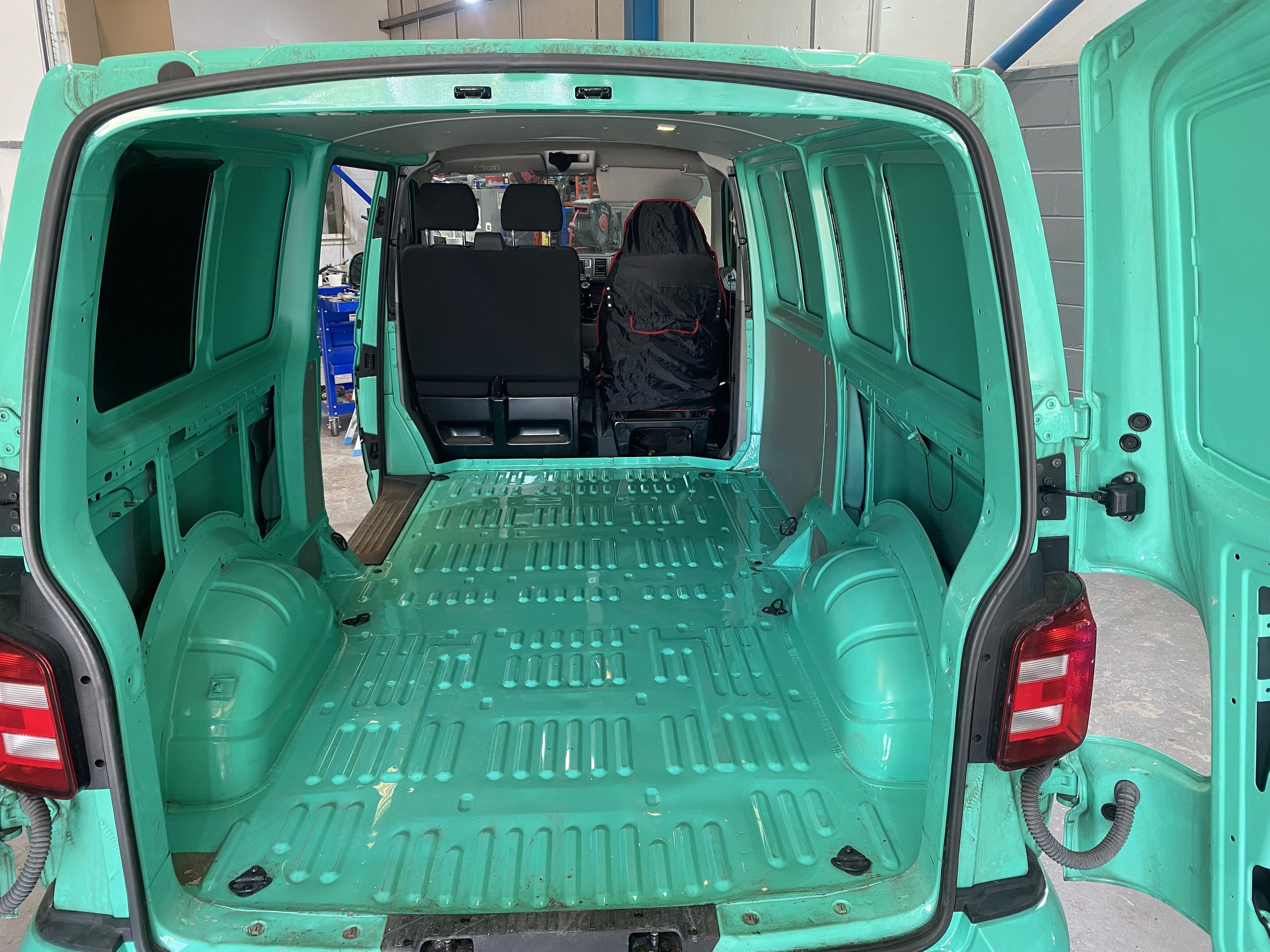 The interior of a small, green vehicle, possibly a van, with the rear door open showing an empty cargo space. The dashboard and two front seats are visible from the back, with the driver's seat covered in a black cloth and the passenger seat covered 