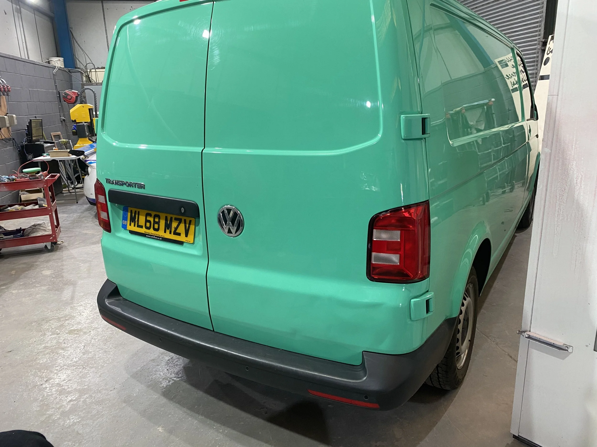 A light green Volkswagen Transporter van inside a garage, with a red tool cart and various tools and equipment in the background.