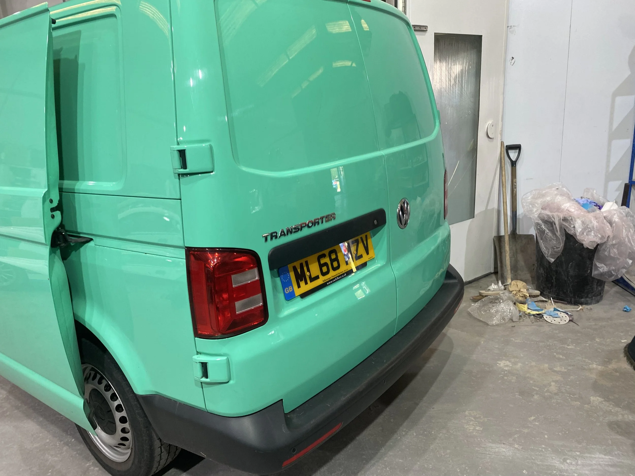 A mint green Volkswagen Transporter van parked inside a garage with a closed rear door. The license plate is visible, and there are cleaning supplies, a shovel, and trash on the floor nearby.