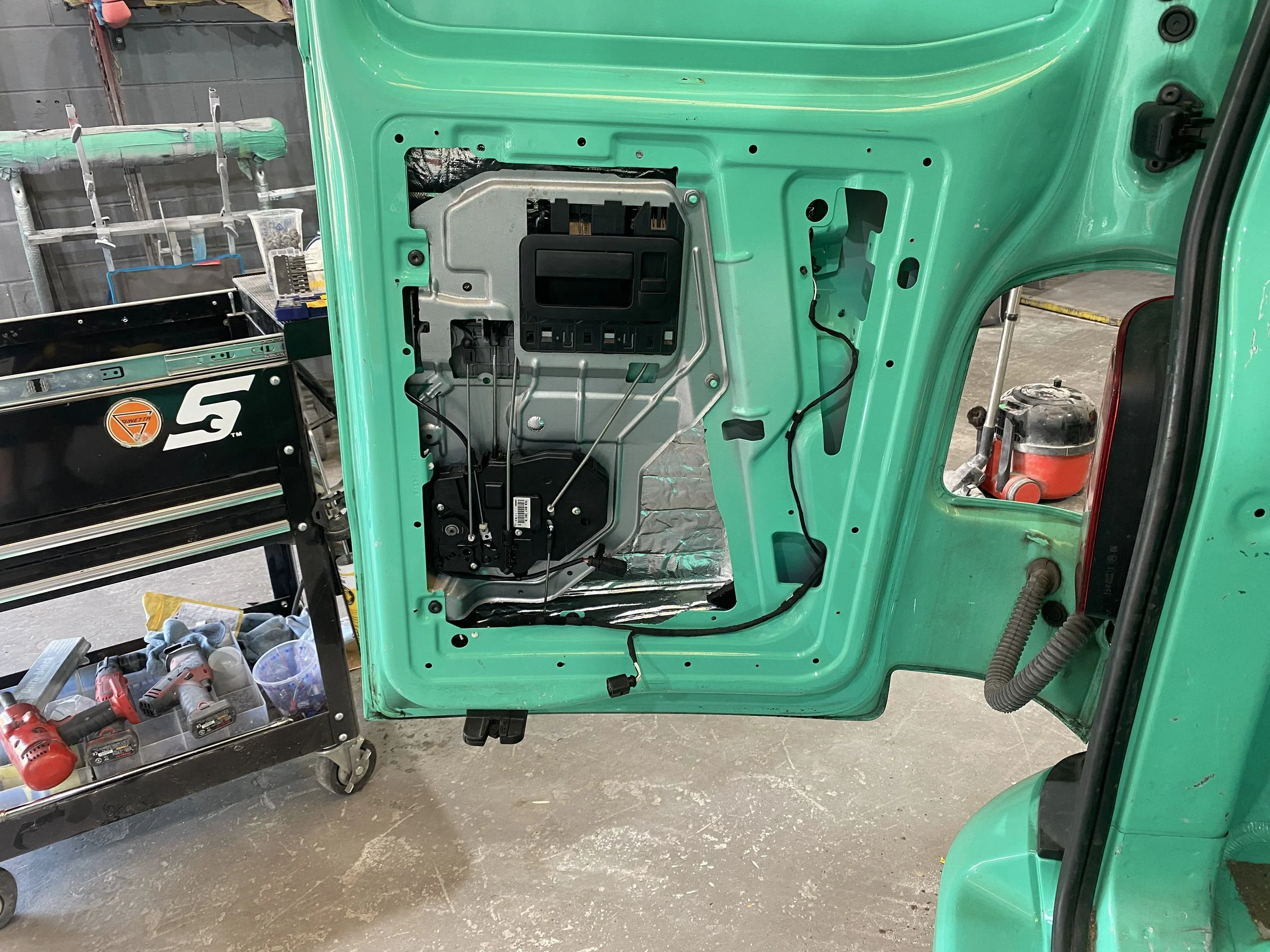 Close-up of a car door with interior panel removed, exposing internal components and wiring, in a workshop setting.
