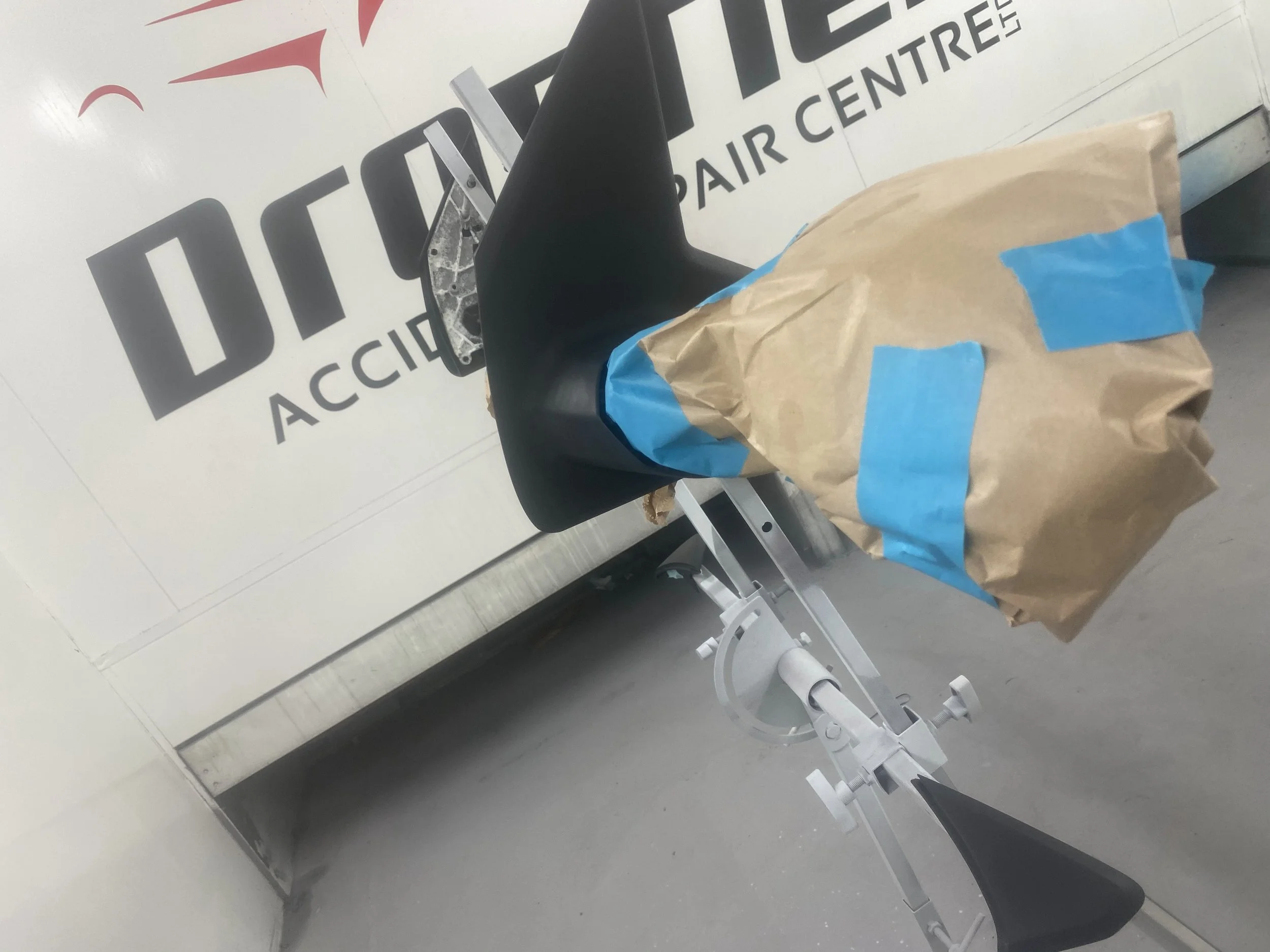 A motorcycle seat covered with a brown paper bag secured with blue painter's tape, attached to a white stand inside an aircraft repair center.