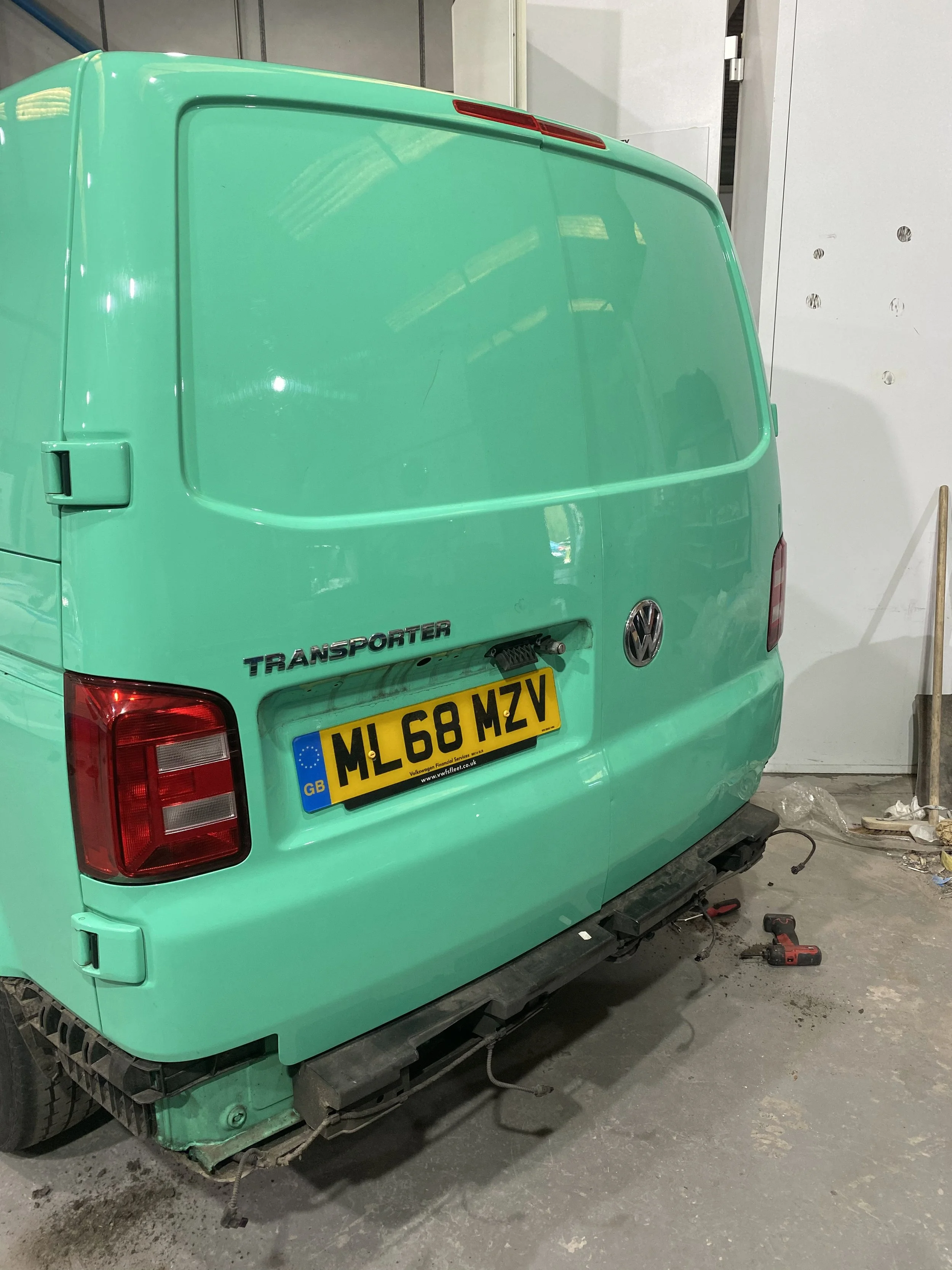 Back of a mint green Volkswagen Transporter van with a UK license plate, parked indoors. Tools and debris are on the floor near the rear bumper.
