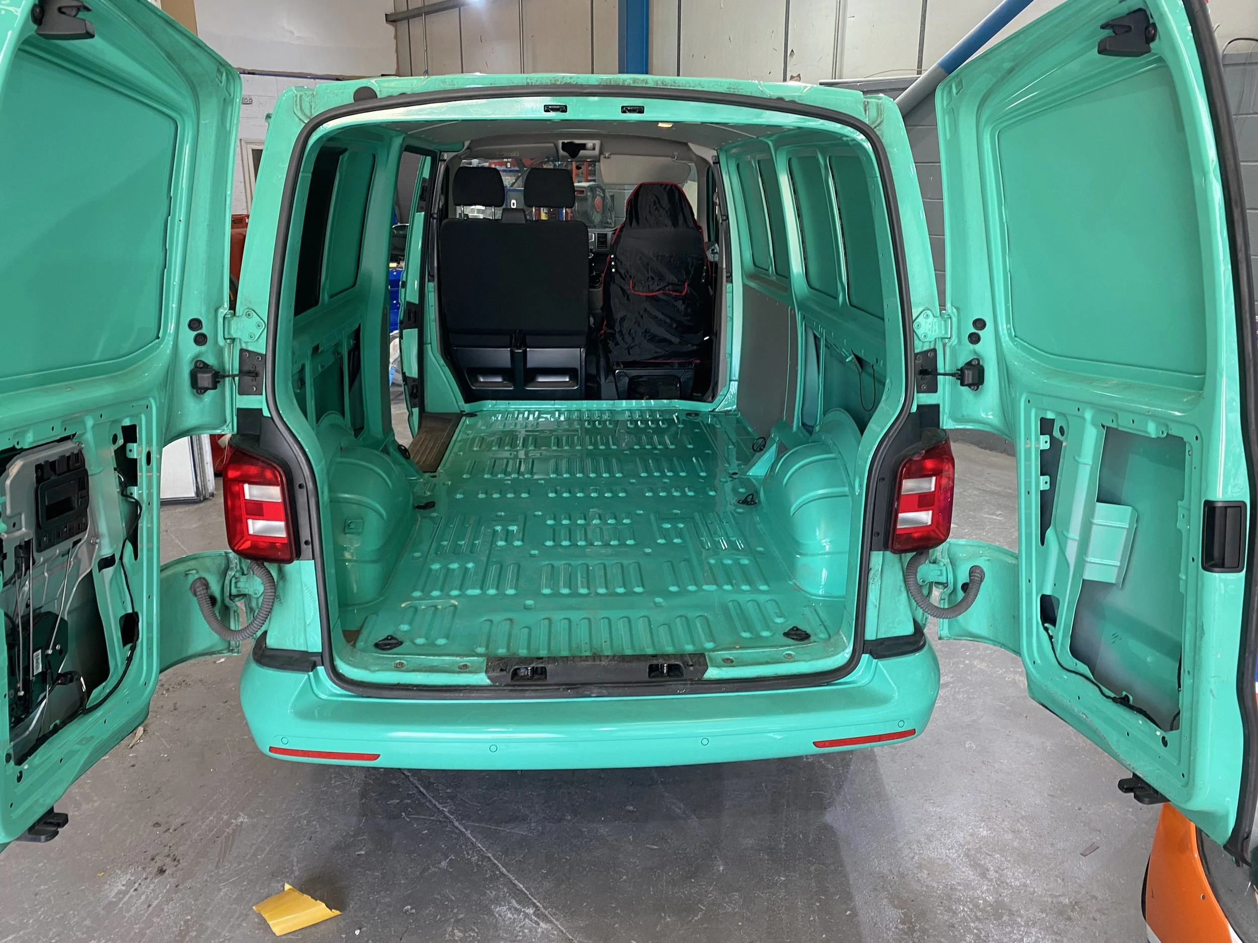 An empty teal cargo van with open rear doors showing the interior, which is stripped down with no seats, exposed metal floor, and stored tools or parts visible at the front cabin.