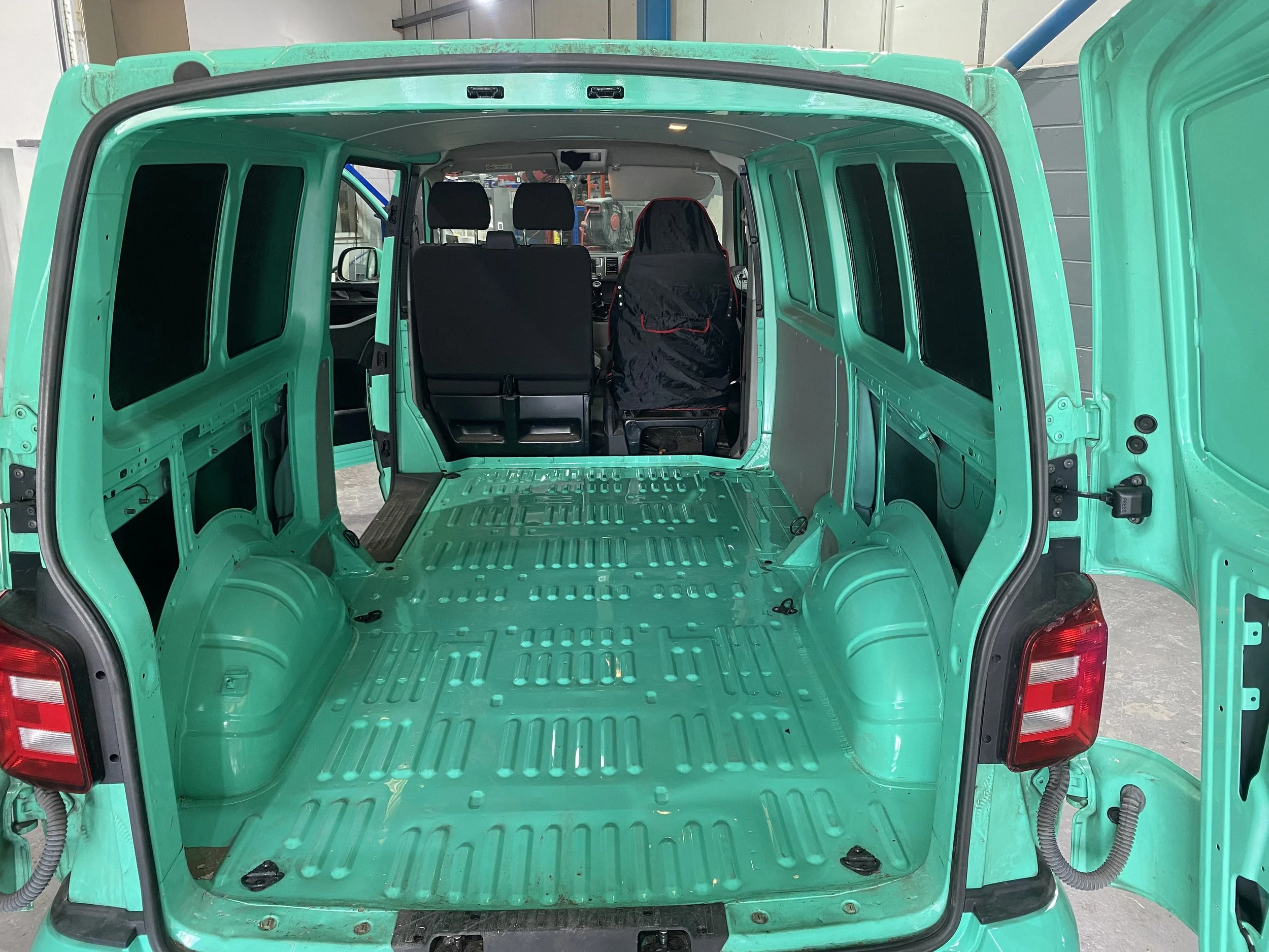 Interior of an empty green van with no seats, showing the metal floor and side panels, and the front two seats visible through the open rear doors.