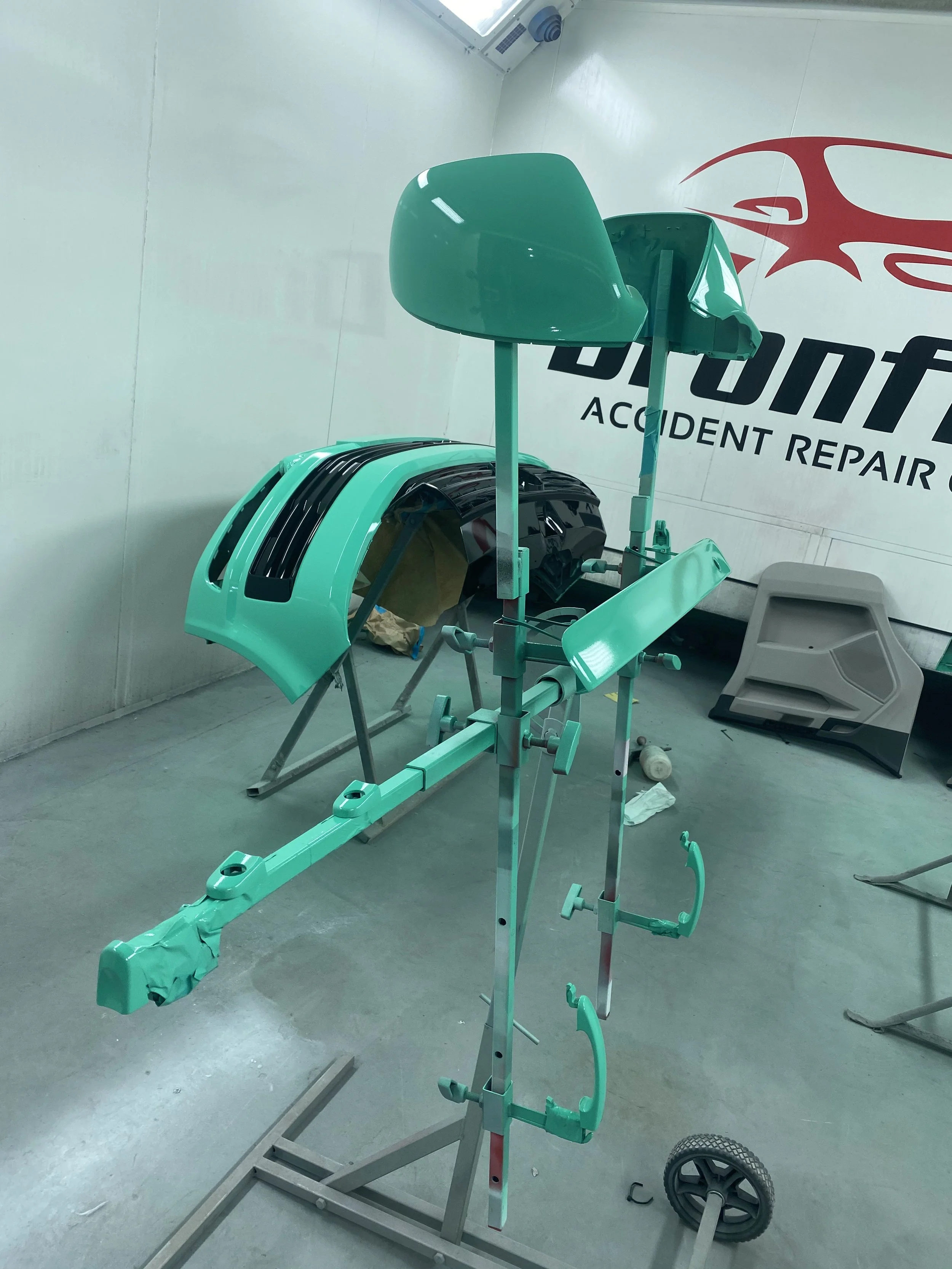 Autobody car parts, including a hood and wing mirror, mounted on a stand in a workshop with a sign that reads 'Accident Repair'.