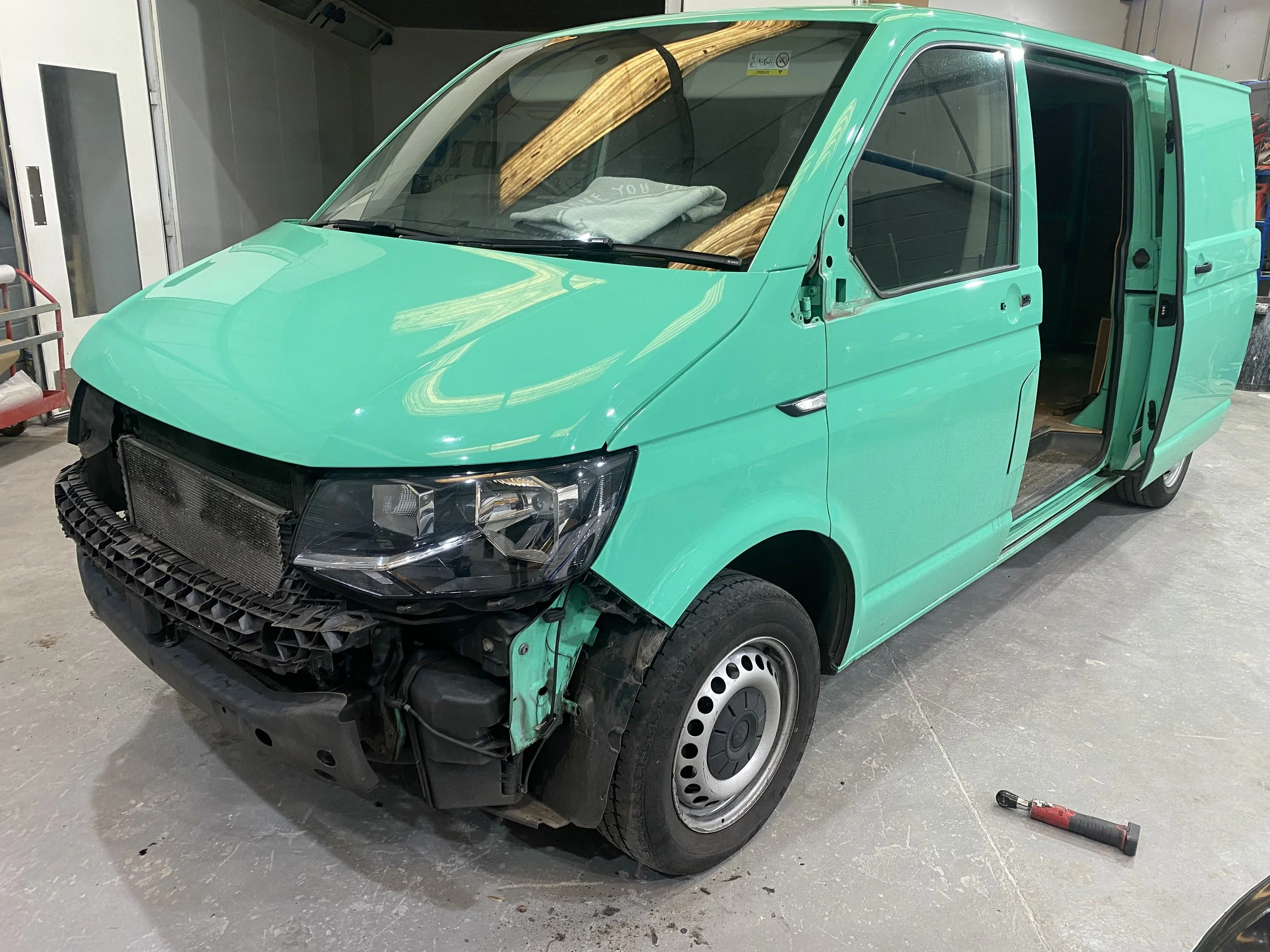 A mint green cargo van with part of the front bumper and grille missing, parked inside a garage or workshop. There is a screwdriver on the ground nearby.