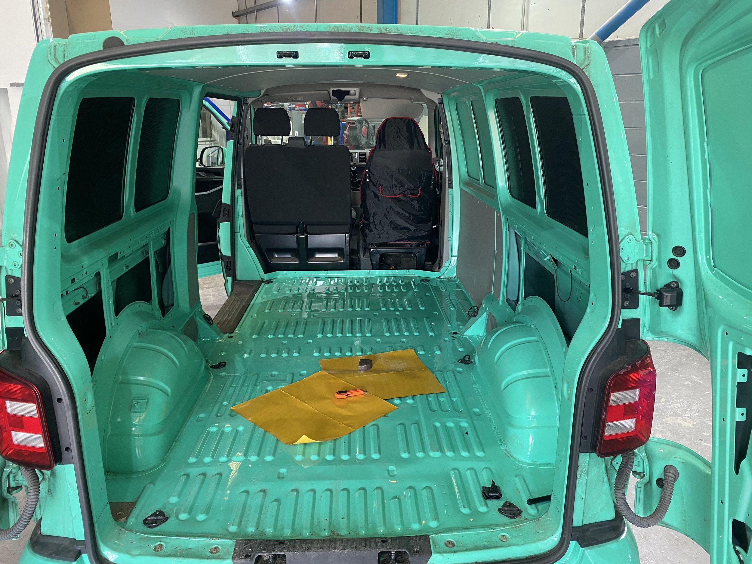 Interior of a mint green cargo van with no seats, showing the metal floor and walls, two seats in the front, and various tools and materials inside.