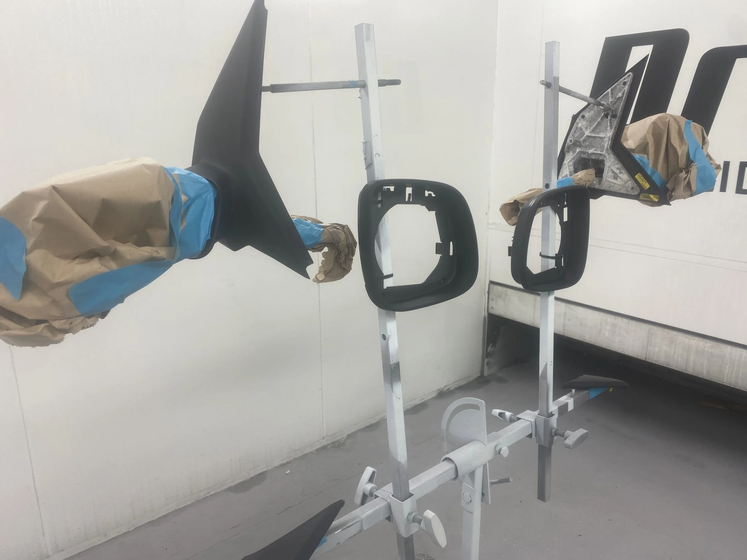 Car parts, including a black rearview mirror and a black trim piece, mounted on a metal stand with a white background.