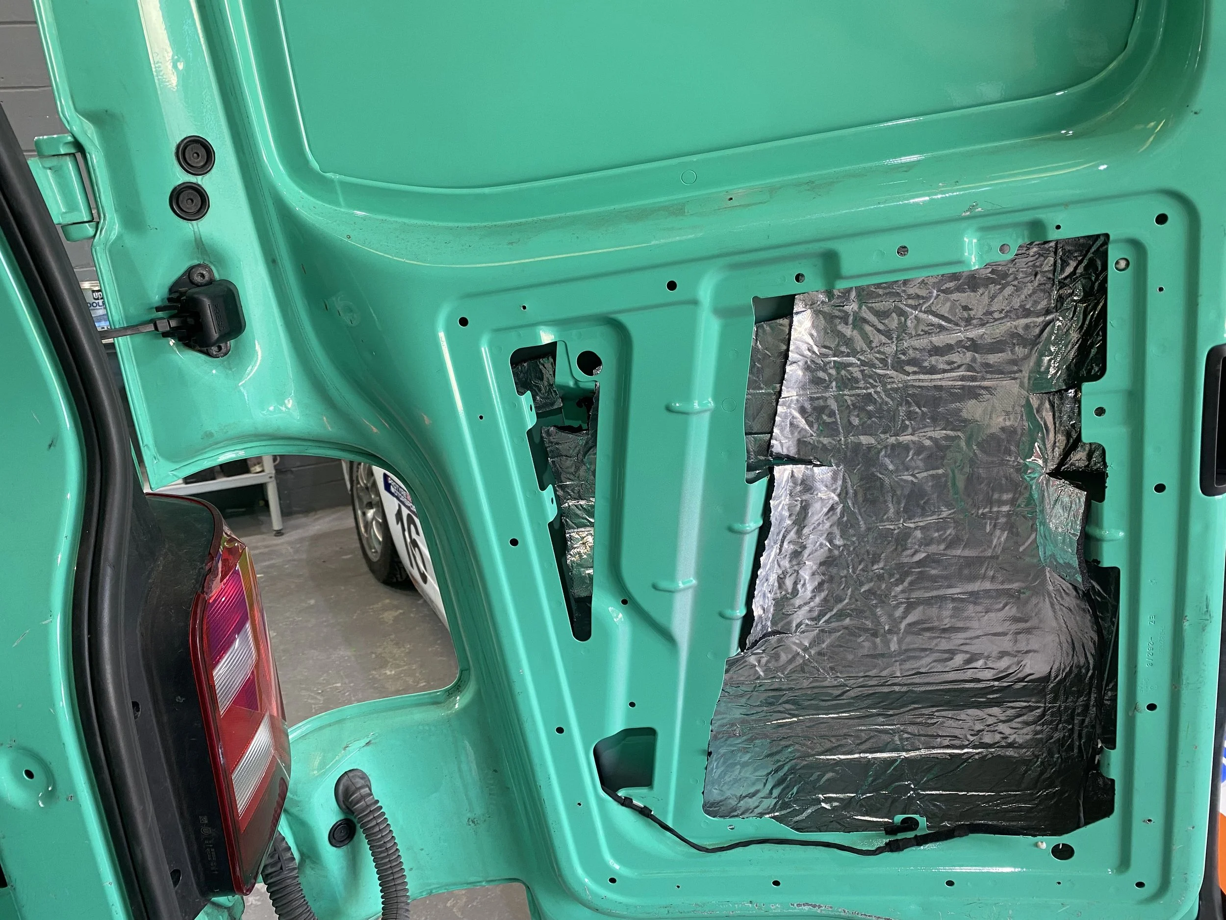 Interior view of a teal-colored vehicle with soundproofing material installed near the rear, showing exposed metal panels and electrical wiring, with a part of the tail light visible.