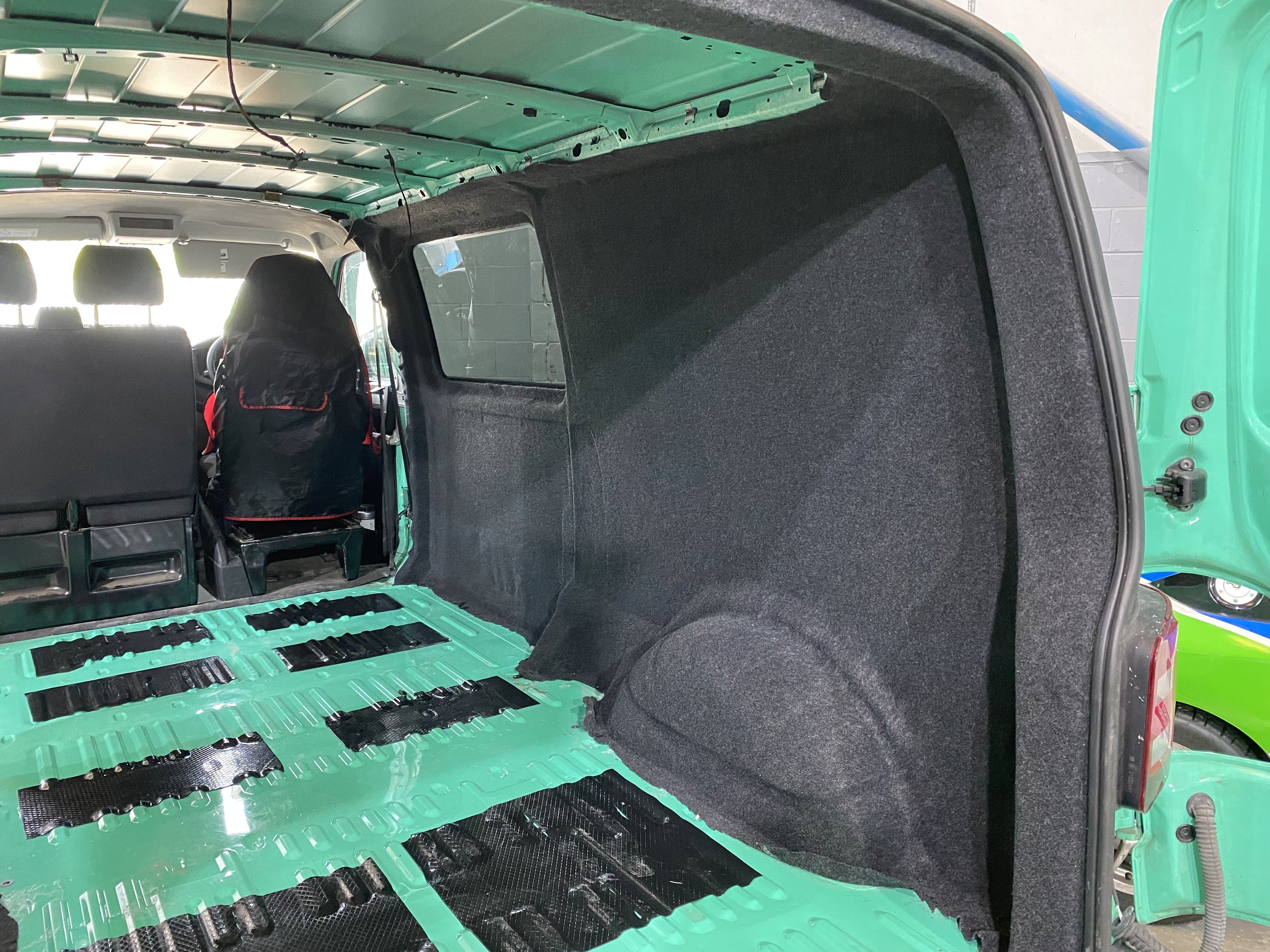 Interior of a vehicle with the rear seats removed, showing the green metal floor with black mounting brackets and the black side panel. The front seats and a window are visible in the background.