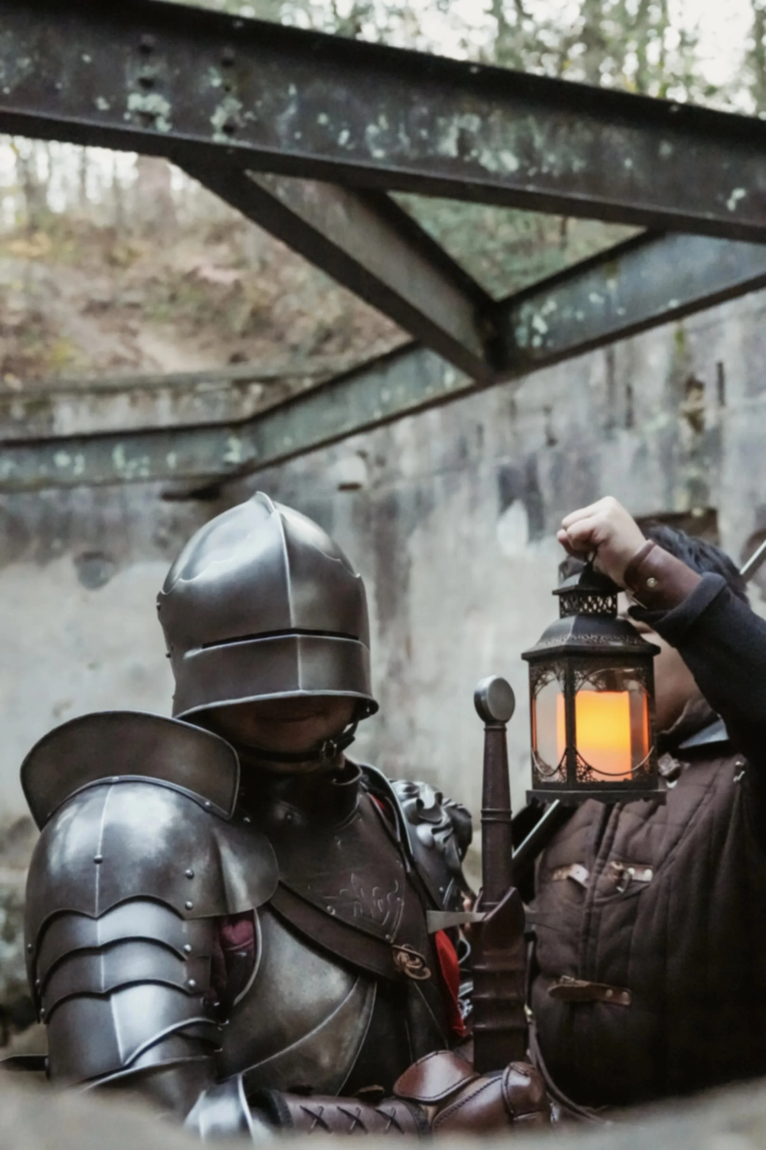A person dressed in medieval-style armor holding a lantern, standing outdoors near a stone wall and trees.