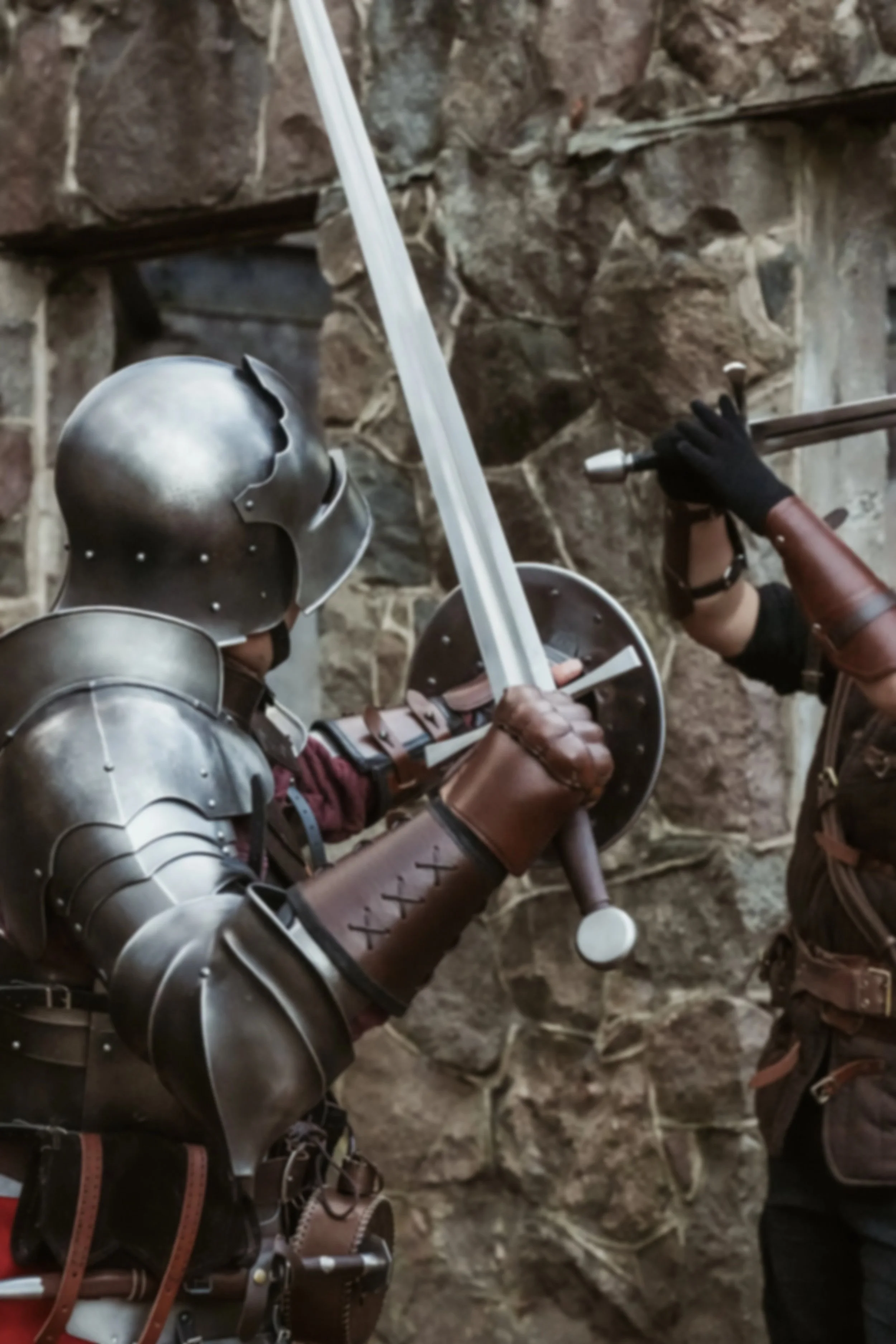 Two individuals dressed as medieval knights practicing sword fighting against a stone wall.