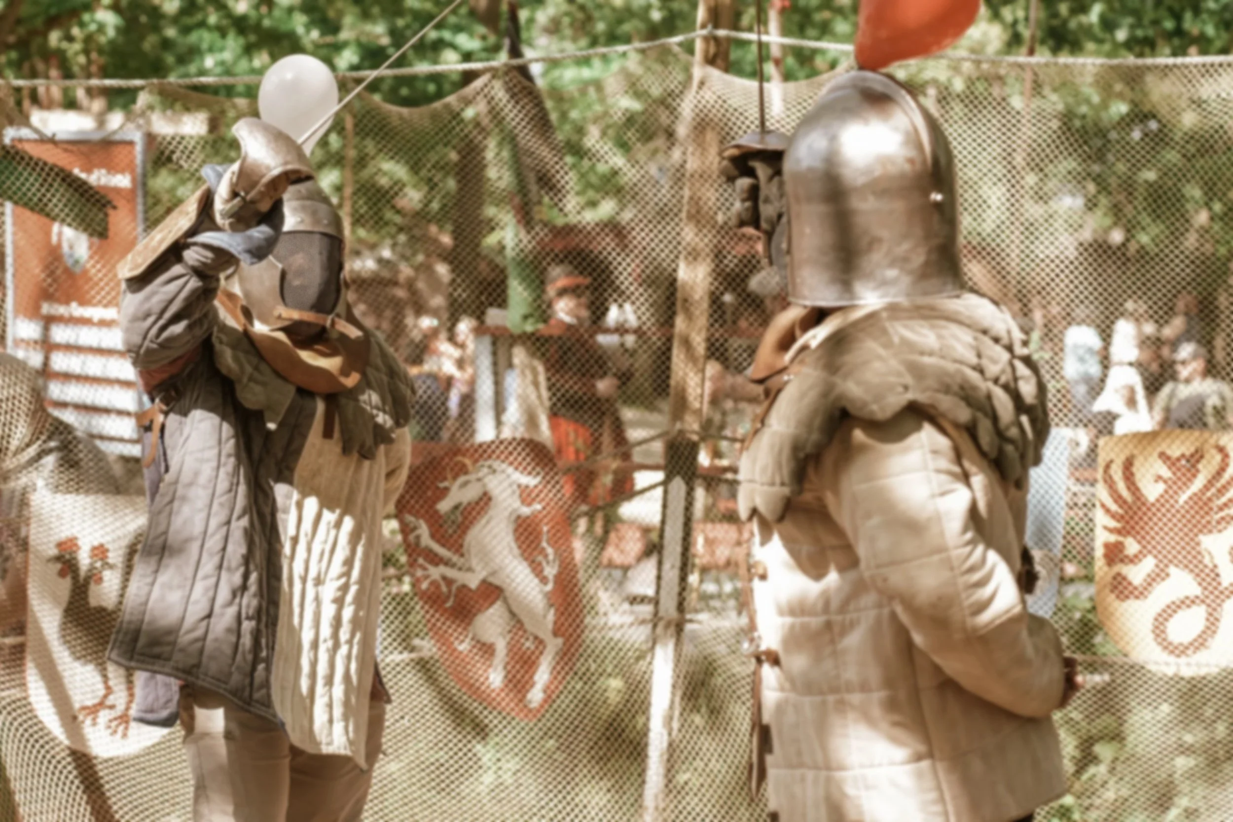 Two individuals dressed as medieval knights with helmets and padded armor engaging in a reenactment or duel at a Renaissance fair or festival, with a backdrop of decorative shields and banners.