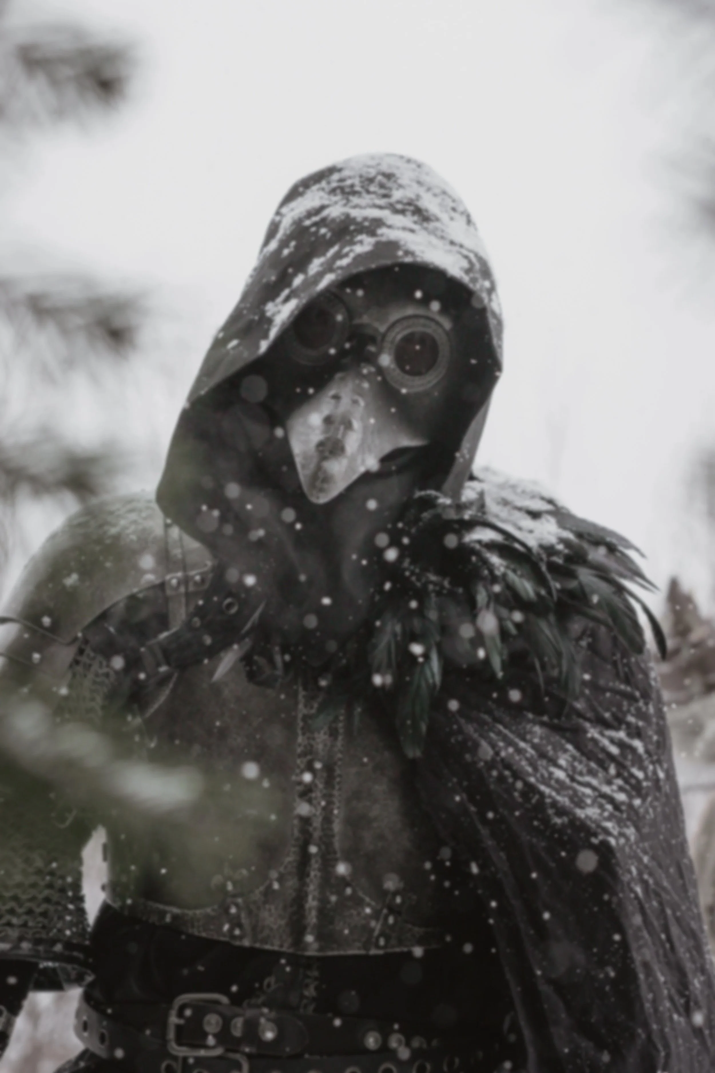 A person dressed in black medieval-style armor with leather and feather details, wearing a plague doctor mask and cloaked in a hood, standing in snowy weather with snowflakes falling.