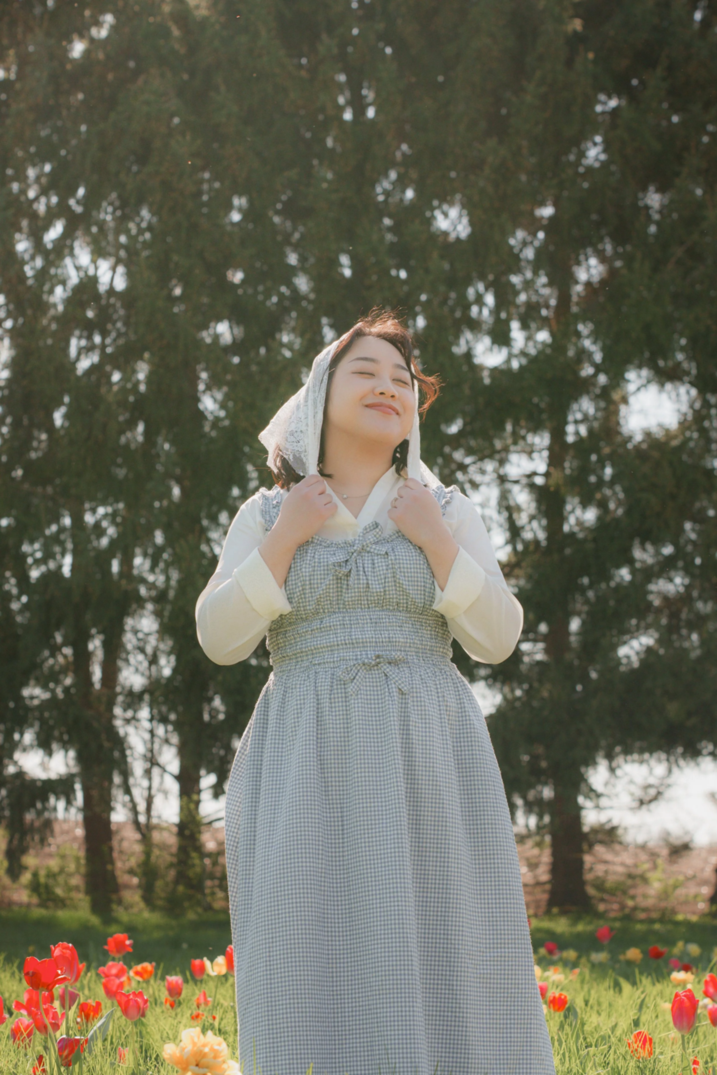 A woman with short brown hair, smiling with her eyes closed, standing outdoors among colorful tulips and green grass, wearing a white headscarf and a blue checkered dress with a white long-sleeve shirt underneath, against a background of tall trees.