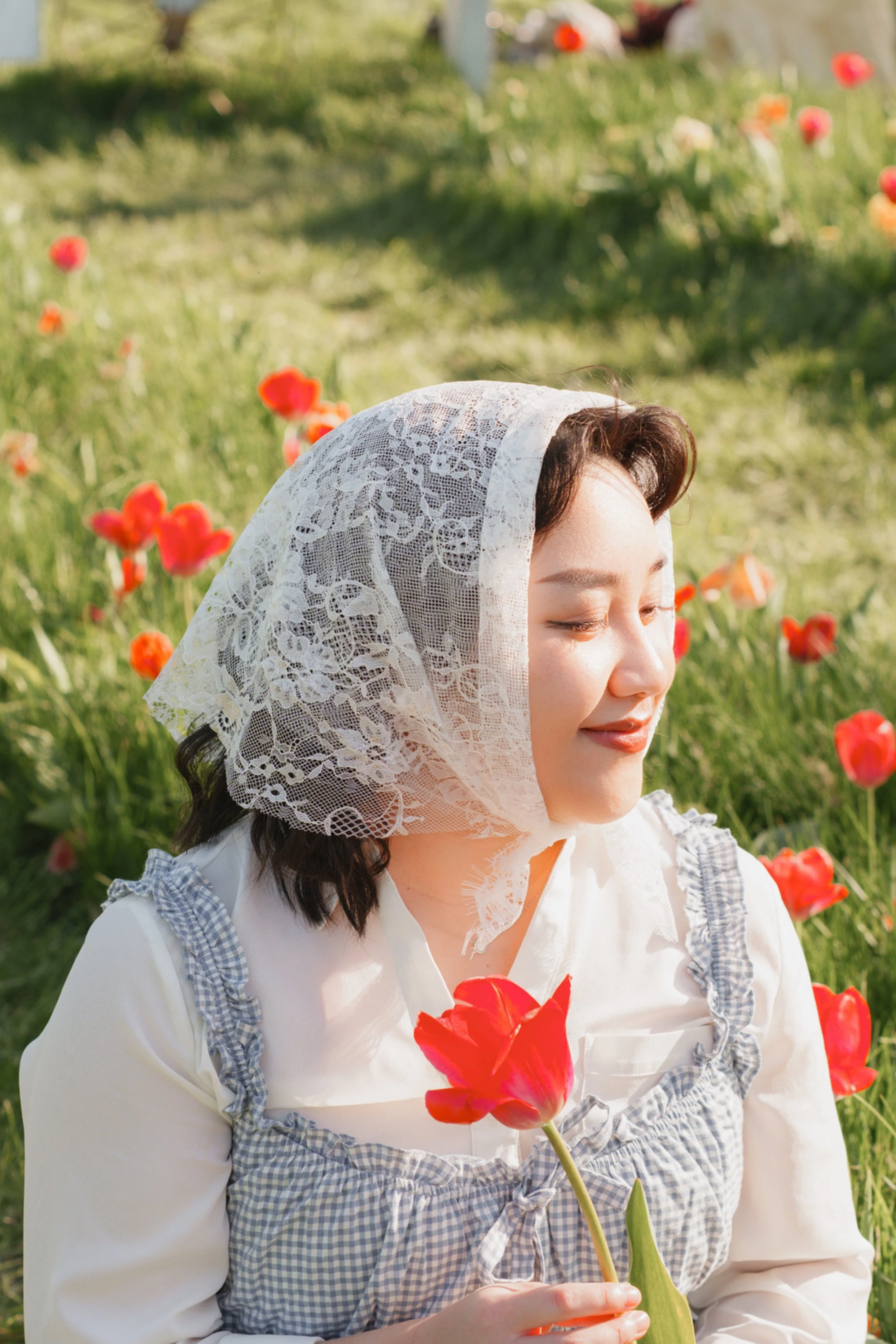A woman wearing a lace headscarf and a white dress with blue checkered ruffles holding a red flower, standing in a field of red tulips with a blurred green background.
