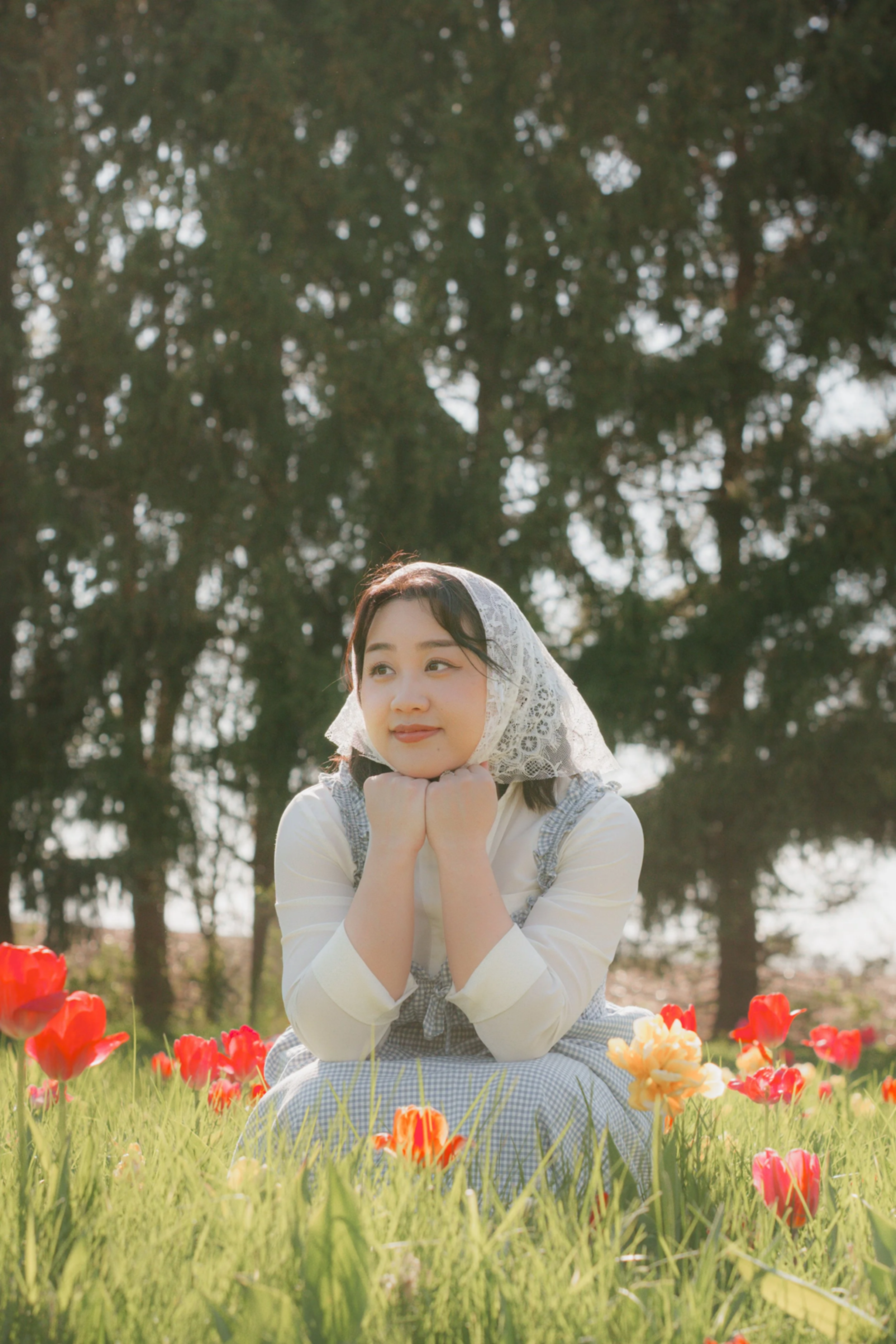 A young woman in a vintage dress and lace head covering sitting on a field of colorful tulips with trees in the background.