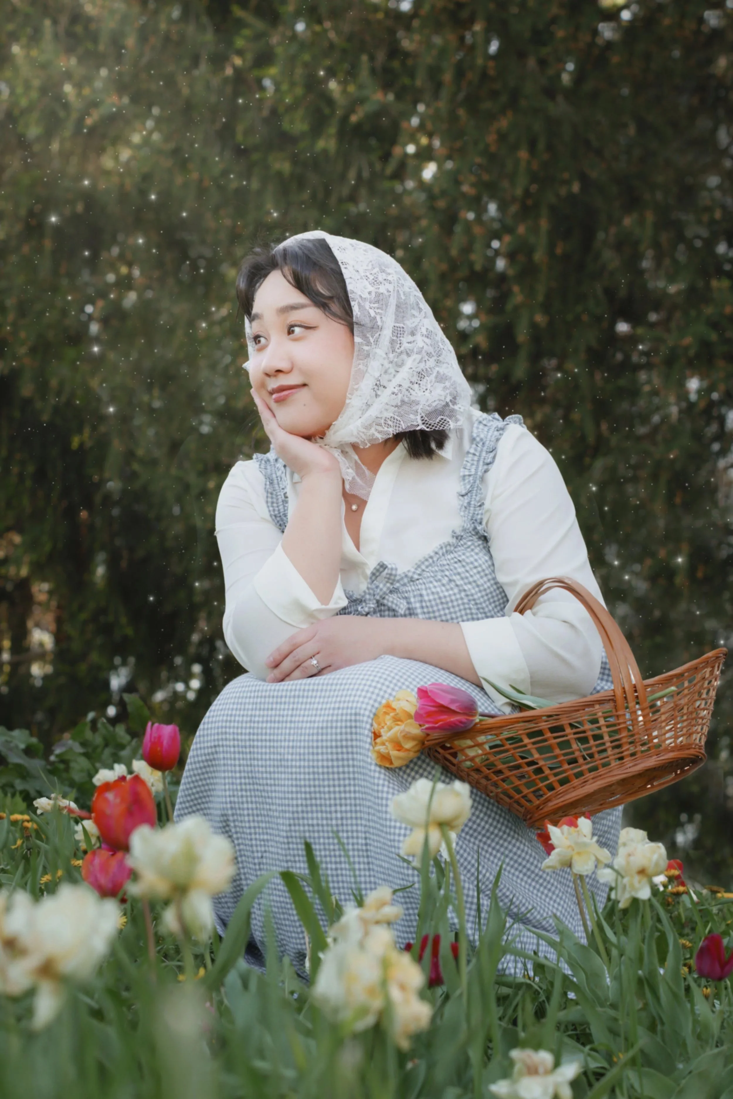 A young woman with short dark hair, wearing a lace bonnet and cheerful expression, is sitting outdoors in a tulip garden during daylight. She is resting her face on her hand, and a basket of colorful tulips is in her lap, with more tulips surrounding