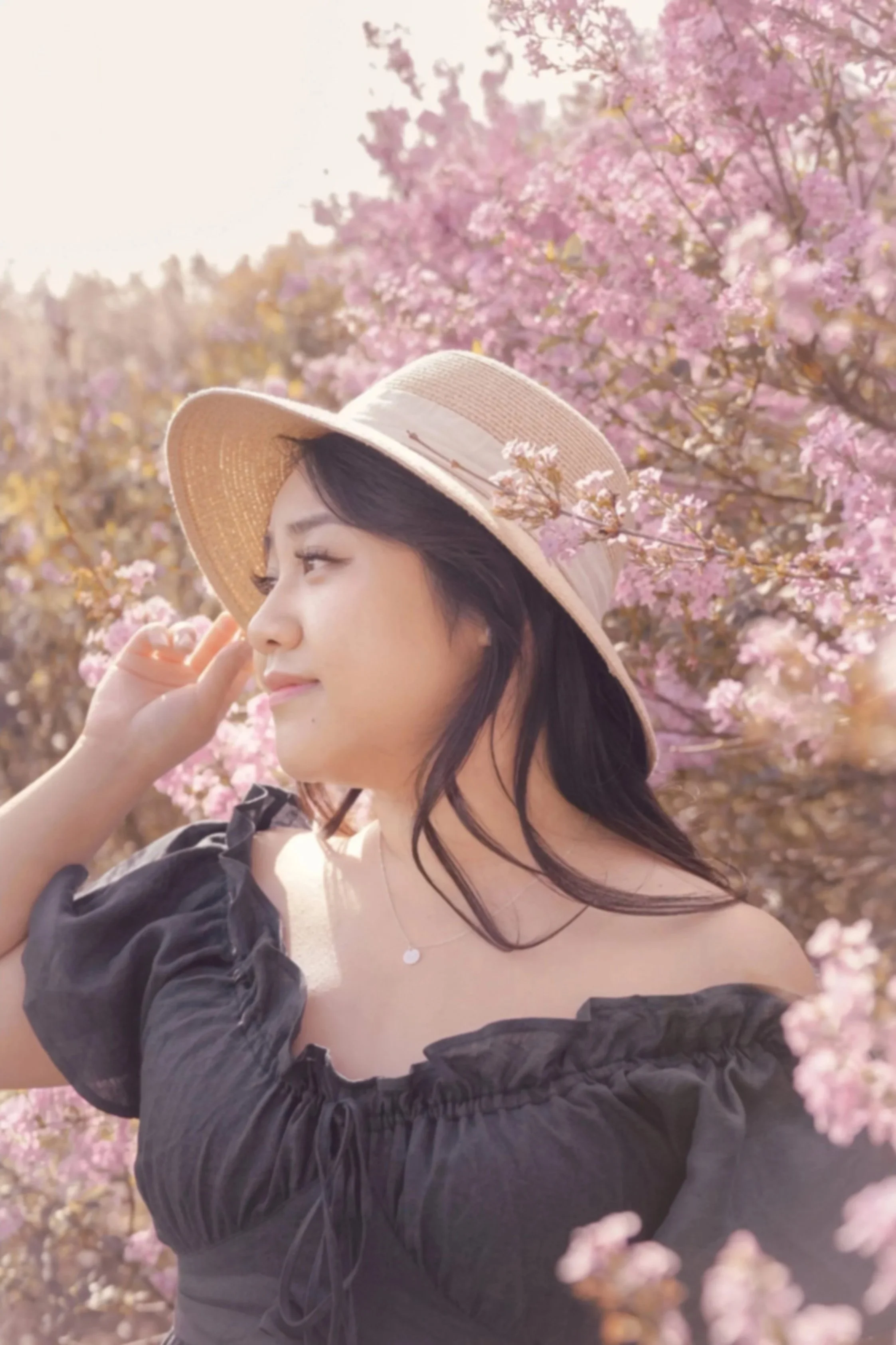 A woman with long dark hair wearing a beige sunhat and black off-shoulder dress standing among pink flowering trees, smiling gently with her eyes closed.