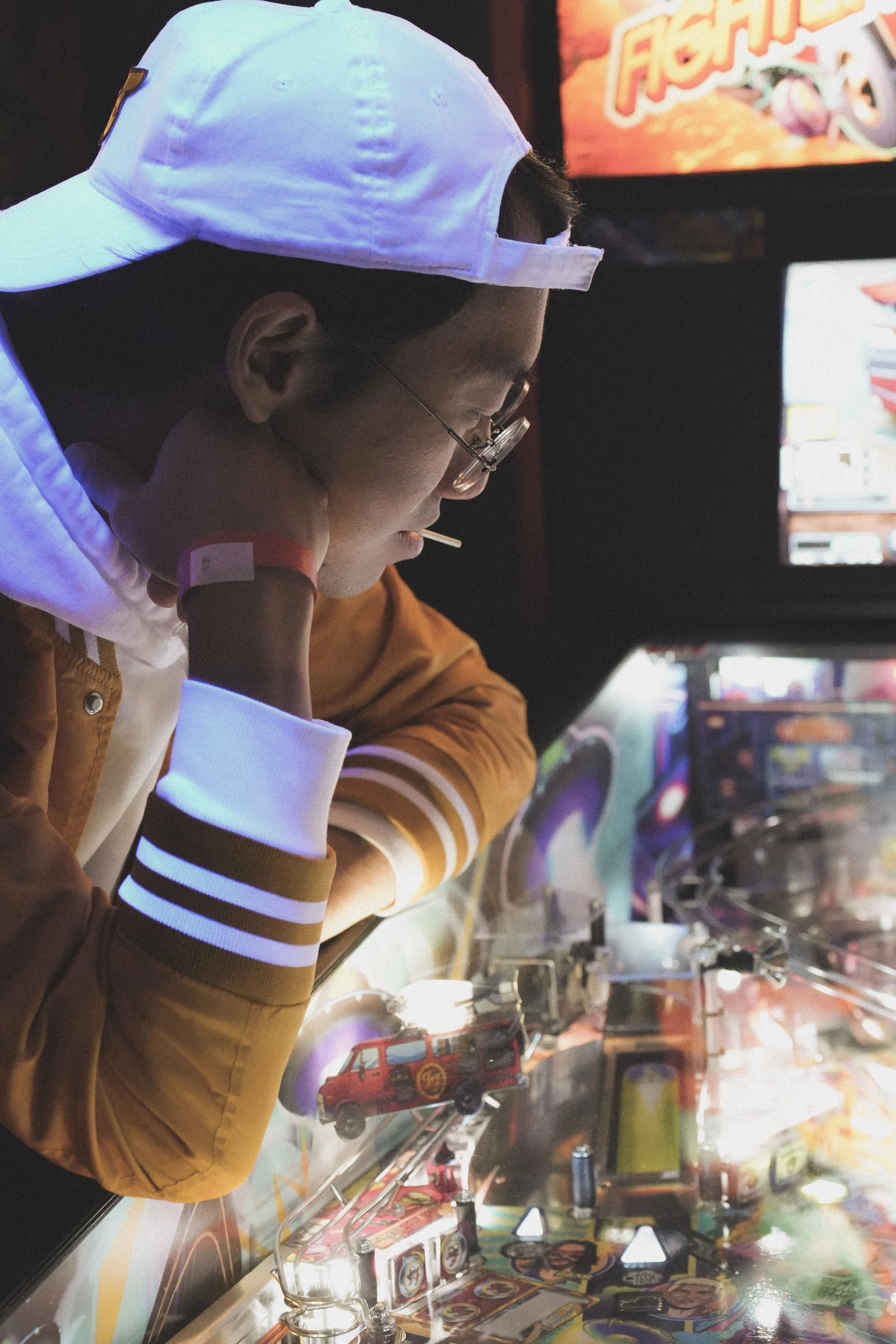 A young man with glasses, wearing a tan and white jacket and a white cap, is playing a pinball machine in an arcade. The arcade has colorful lights, and there is a display screen showing the game.