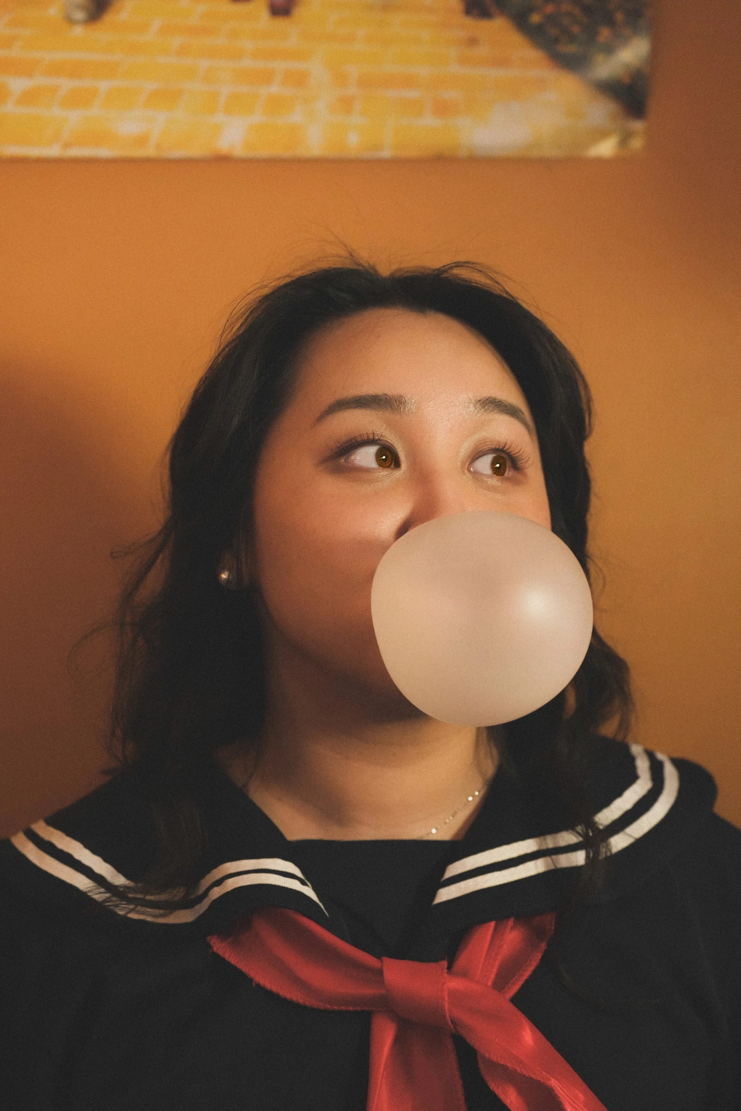 A woman with dark hair and light skin blowing a bubble with bubblegum, wearing a black top with white stripes and a red bow, against an orange background with a partially visible piece of artwork.