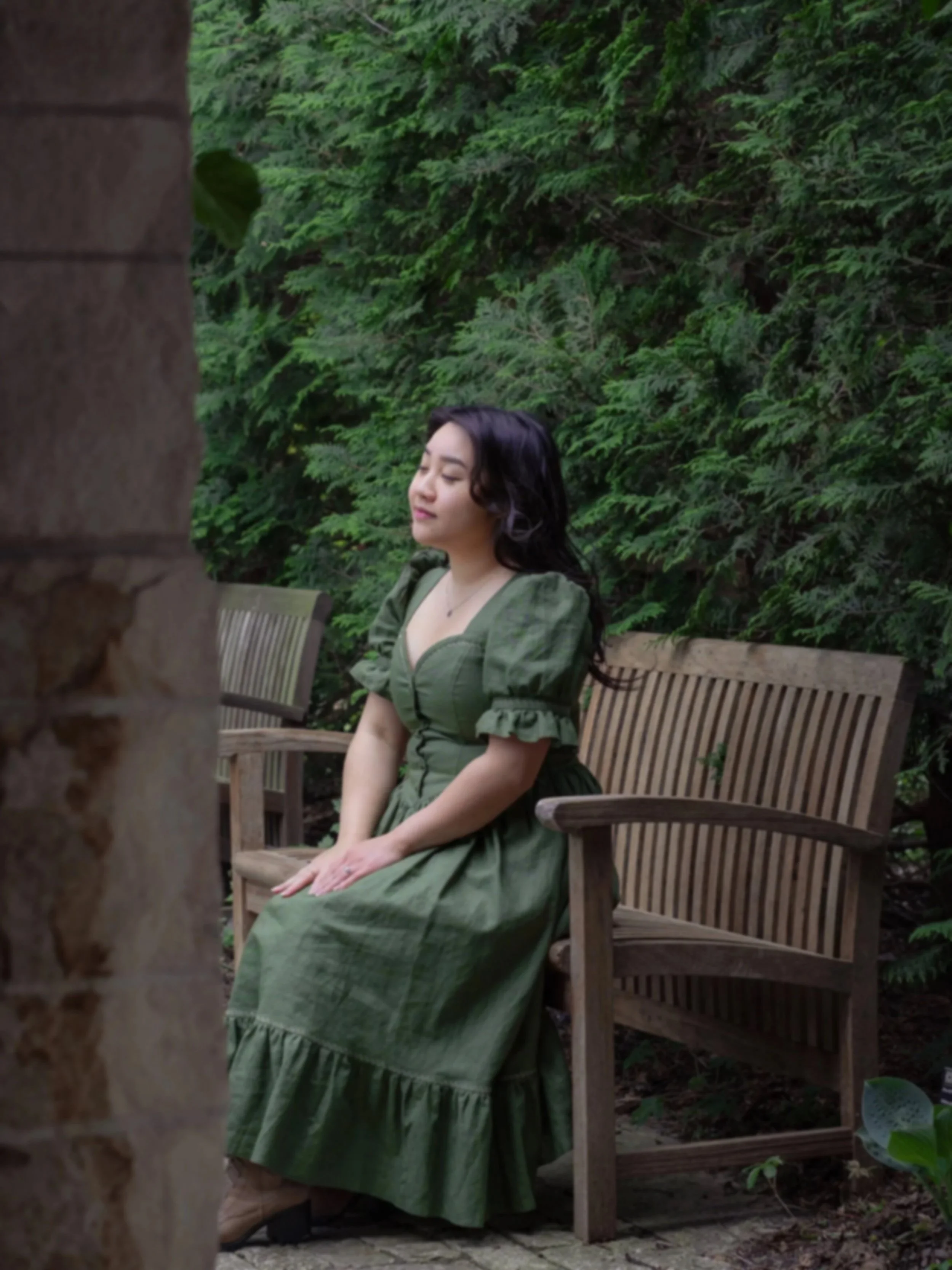 A woman in a green dress sitting on a wooden bench in a garden surrounded by lush greenery.