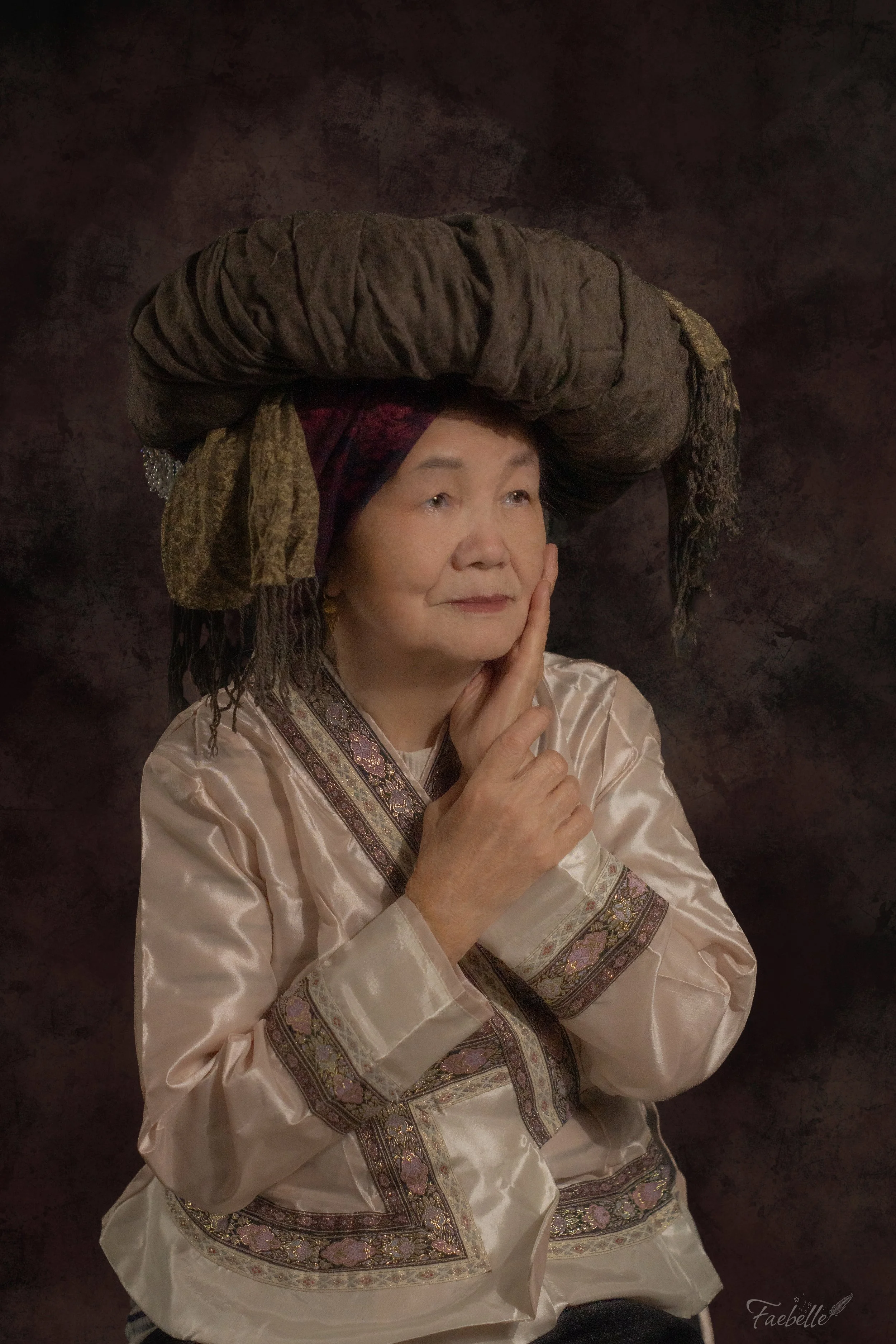 An elderly woman with light complexion, short gray hair, and a gentle smile, wearing an elegant cream-colored traditional dress with intricate embroidered patterns in gold, pink, and purple, and a large headpiece made of layered, fabric-rolled materi