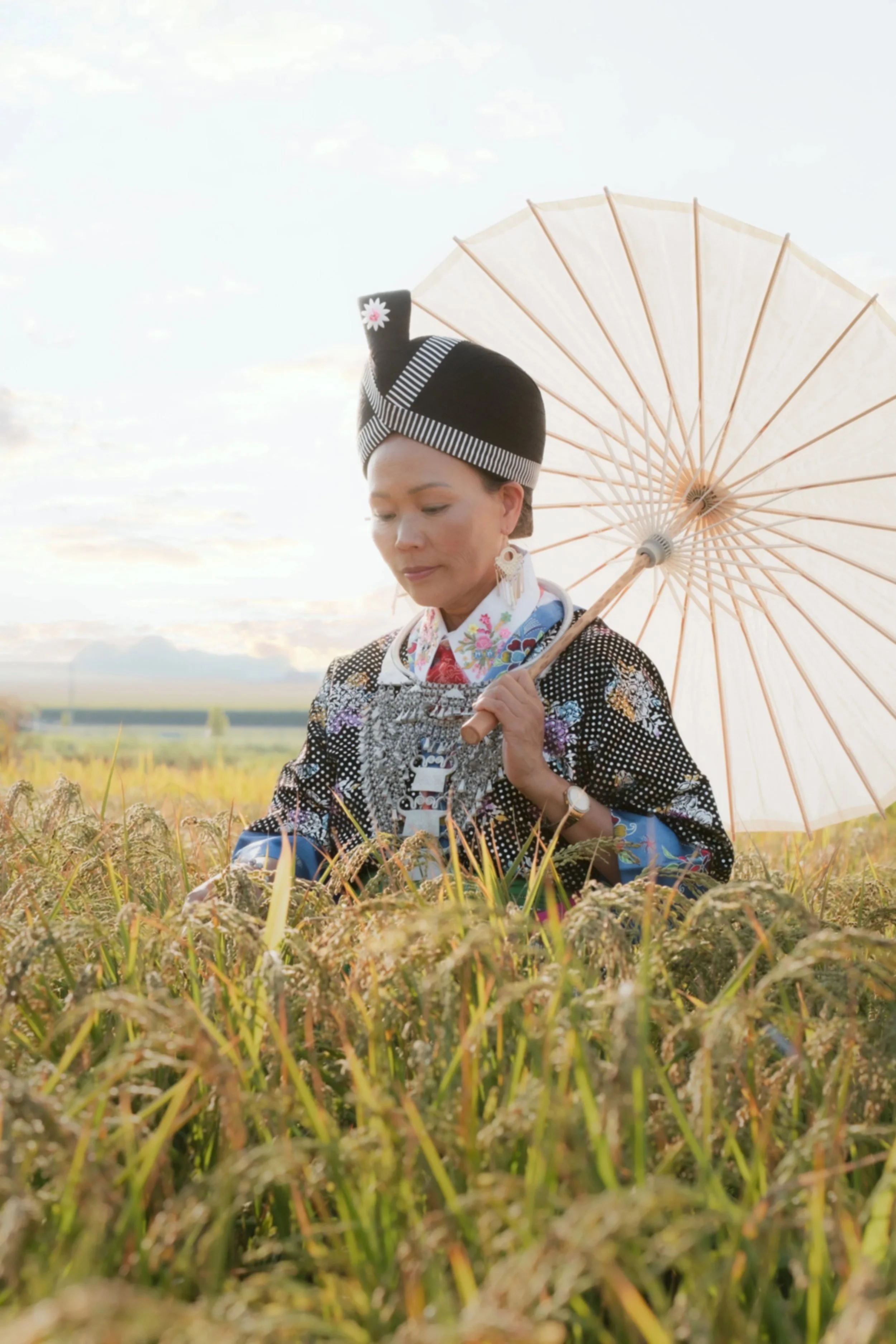 A woman in traditional Hmong attire stands in a rice field, holding a parasol and looking down at the rice plants.