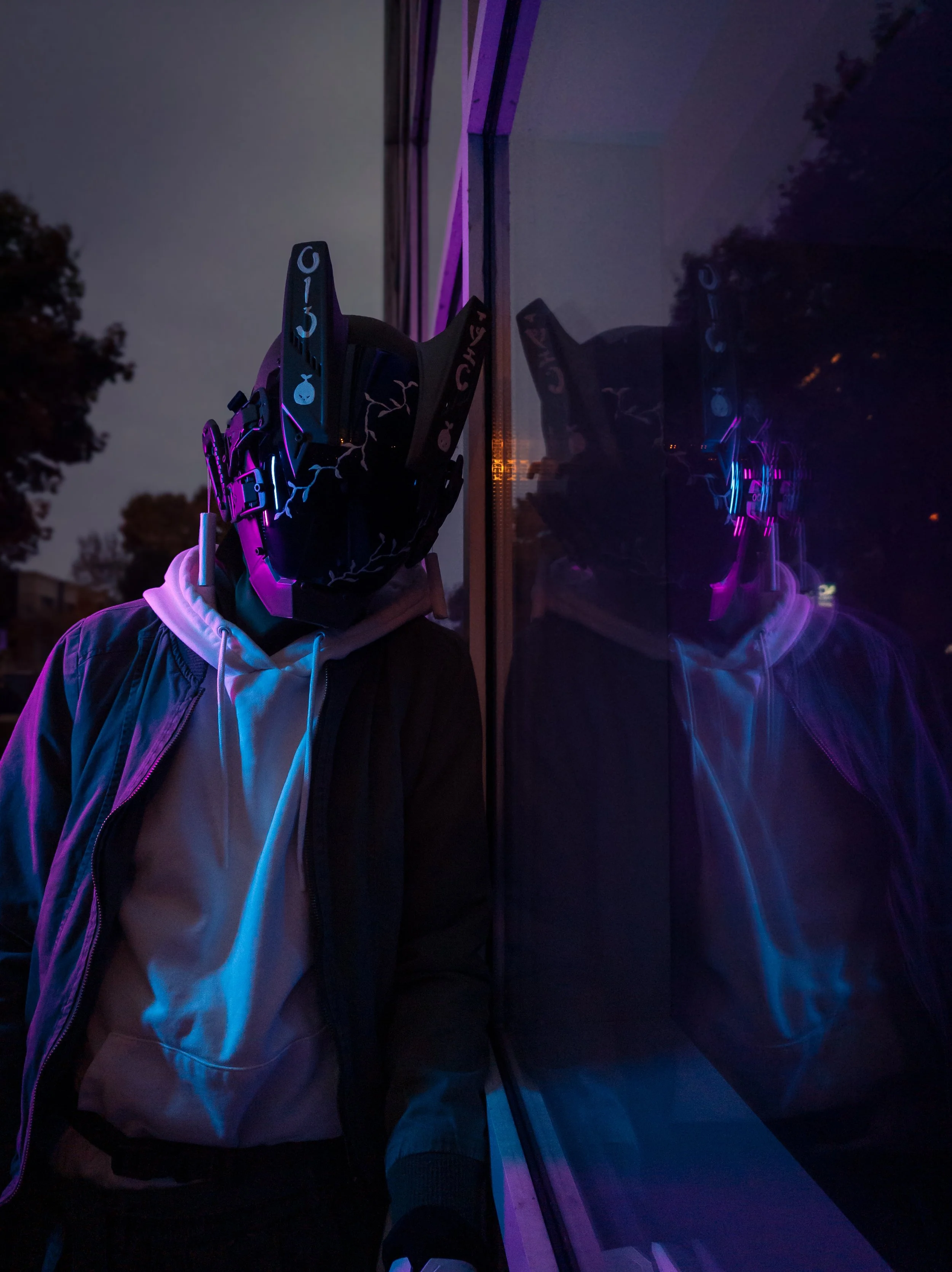 Person wearing a ski or snowboard helmet and jacket, standing outside at night, with their reflection visible on a glass surface.