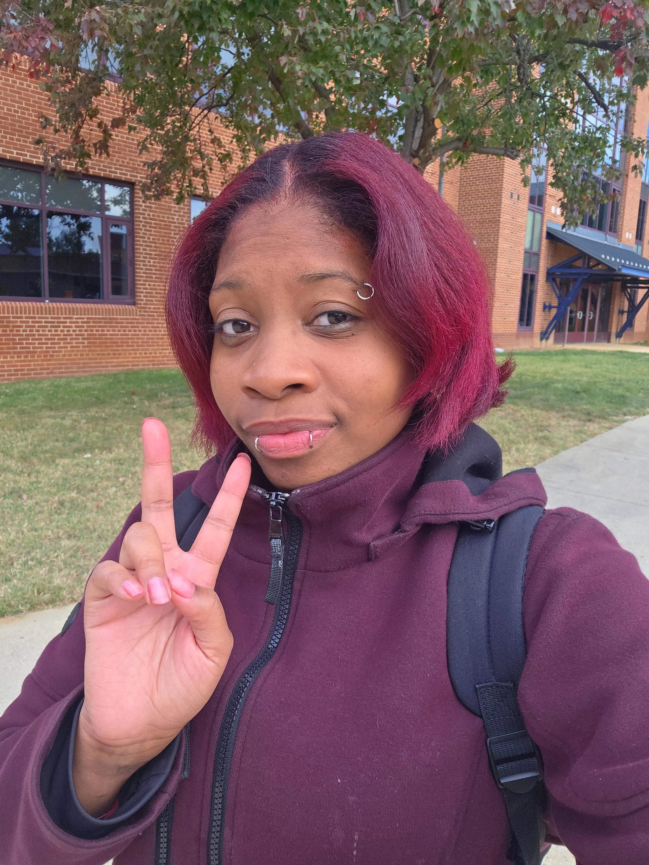 어깨 길이의 보라색 염색 머리를 한 여성이 벽돌 건물과 나무 앞에서 브이 사인을 하고 있습니다.