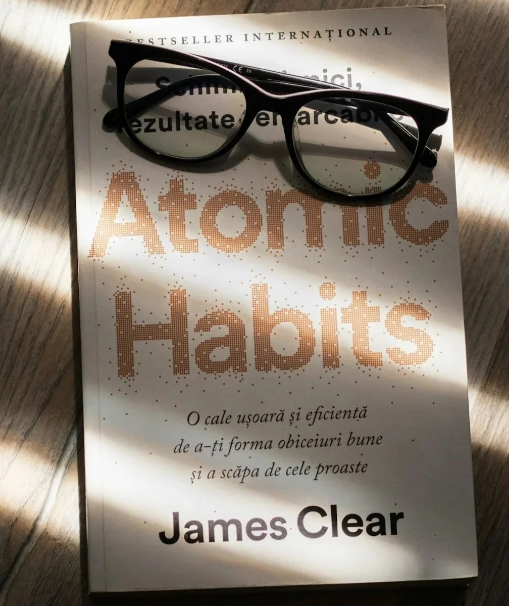 A book titled "Atomic Habits" by James Clear with a pair of black eyeglasses resting on top of it. Part of the book cover is illuminated by sunlight and some shadow.