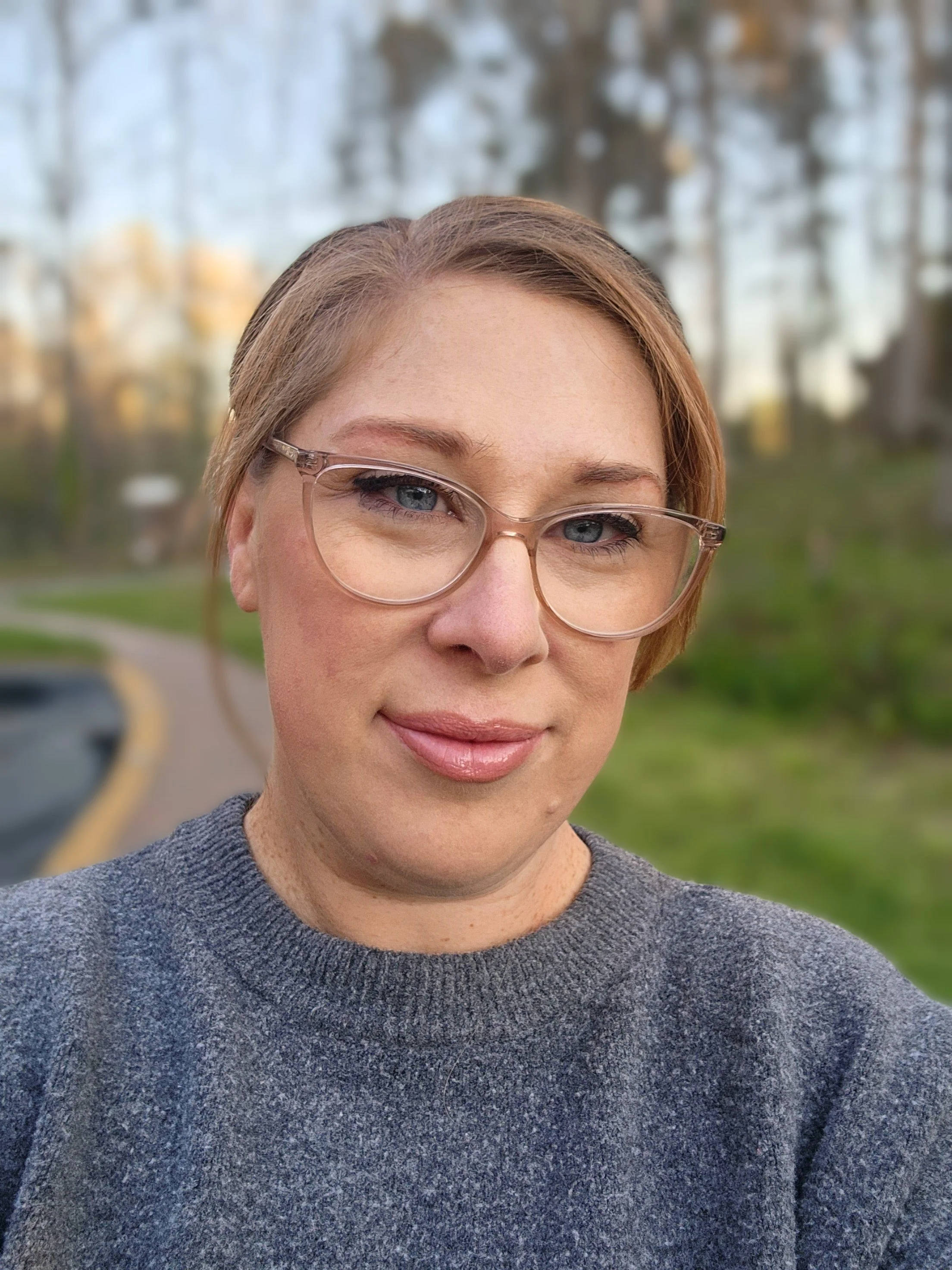 Close-up of a woman with short, reddish-brown hair, wearing clear eyeglasses and a gray sweater, standing outdoors with a blurred background of trees and a park pathway.