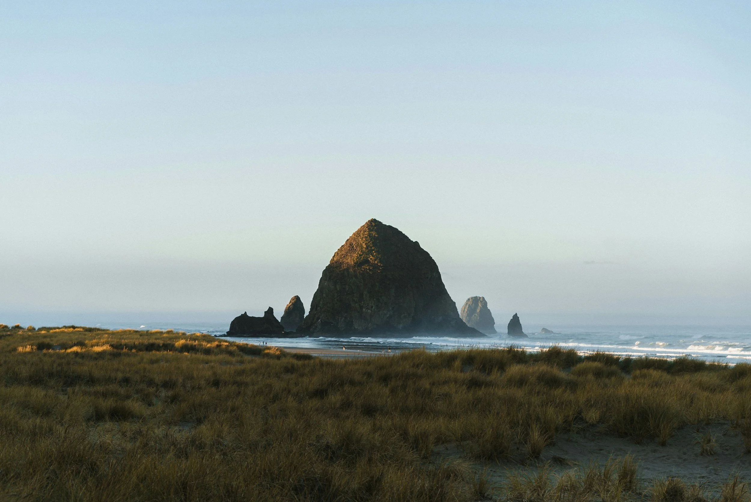 A large sea stack formation rising from the ocean near a sandy beach with grassy dunes in the foreground, under a clear sky.
