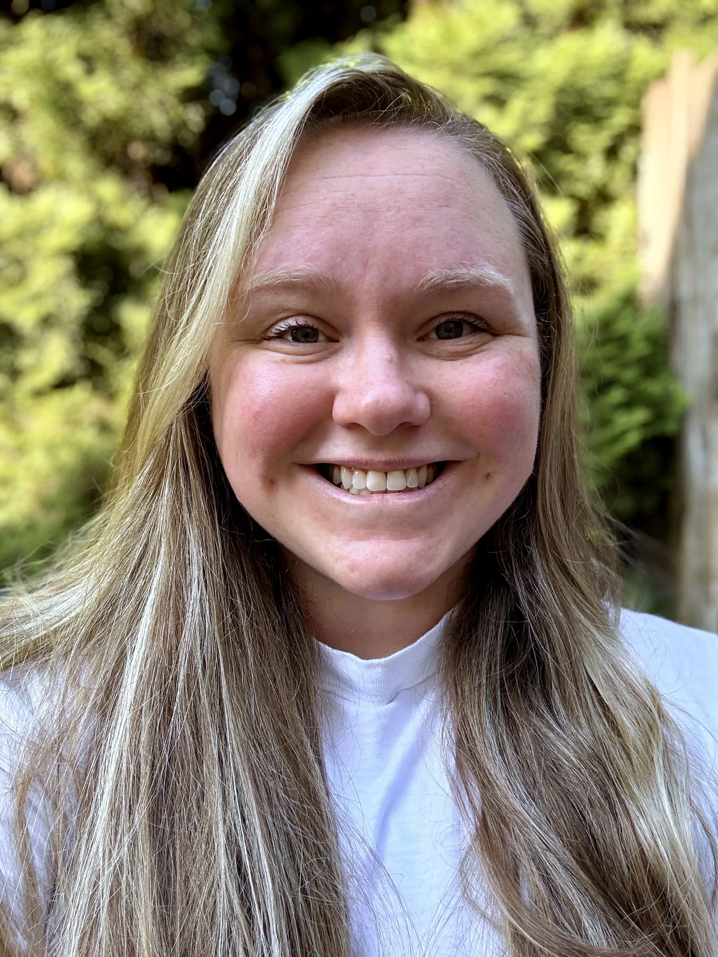Close-up of a smiling woman with long blonde hair, outdoors with greenery in the background.