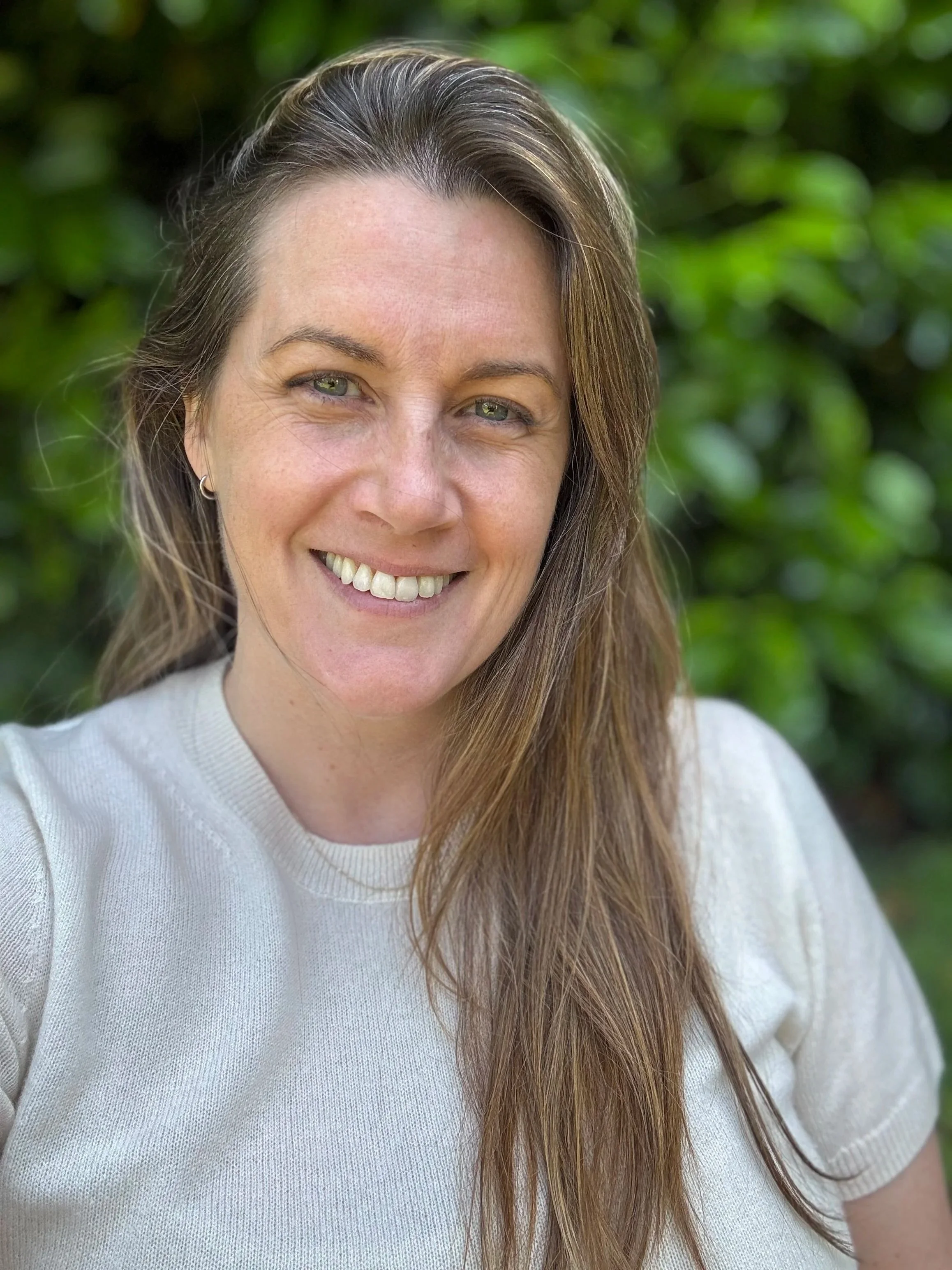 A woman with long brown hair, smiling, wearing a white sweater, outdoors with green foliage in the background.