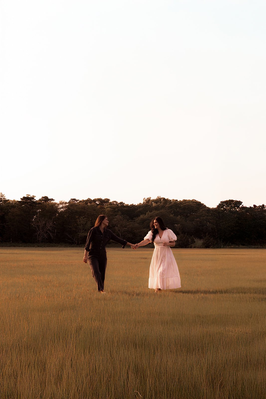 Two women laughing together barefoot on the beach during a Cape Cod anniversary session, candid LGBTQ+ photography