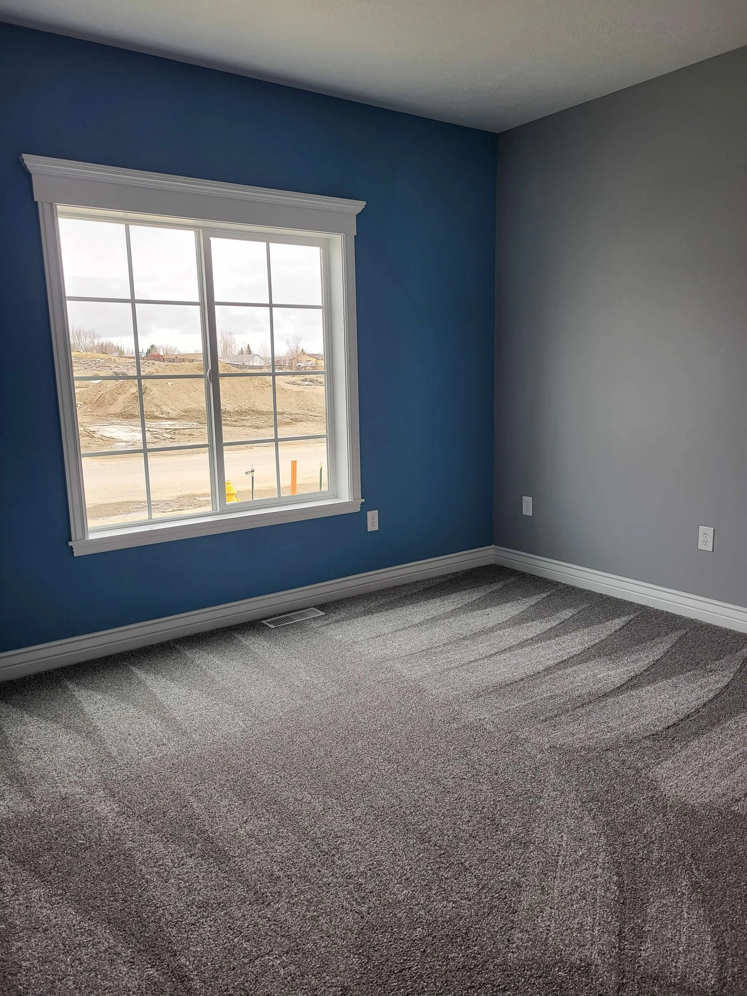 Empty room with carpet flooring, blue accent wall, and a large window showing an outdoor construction site.