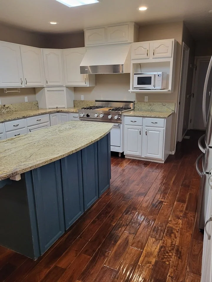 A kitchen with white cabinets, a stainless steel stove with an oven, a microwave in a corner, granite countertops, a blue kitchen island, and wooden flooring.