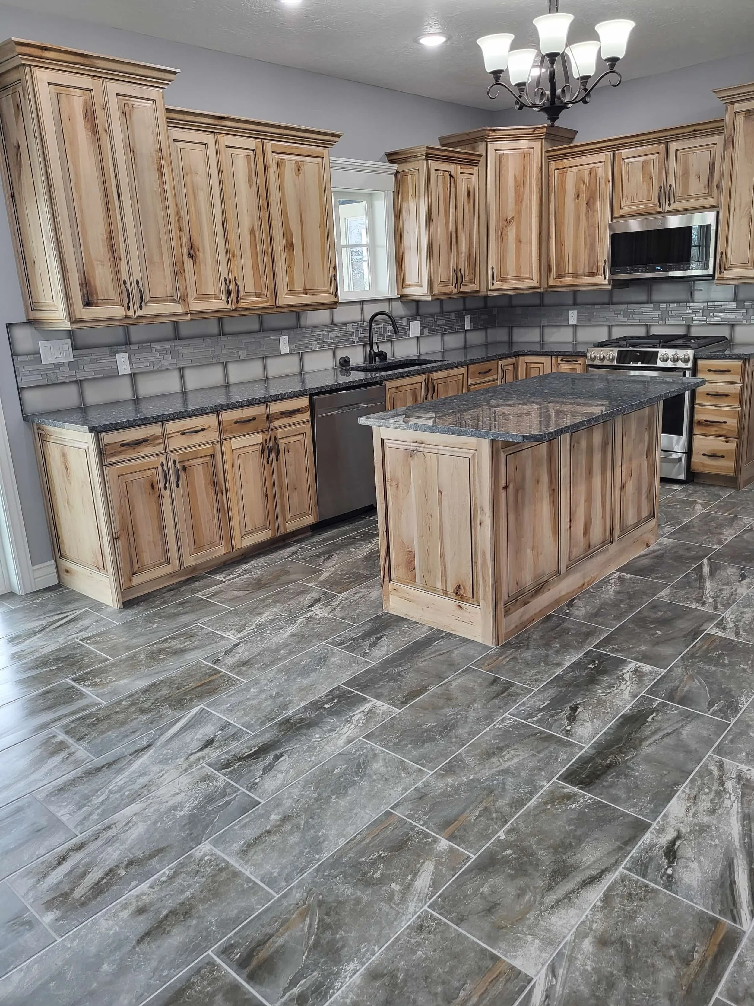 Kitchen with wood cabinetry, granite countertops, a small island, stainless steel appliances, gray tile backsplash, and patterned floor tiles.