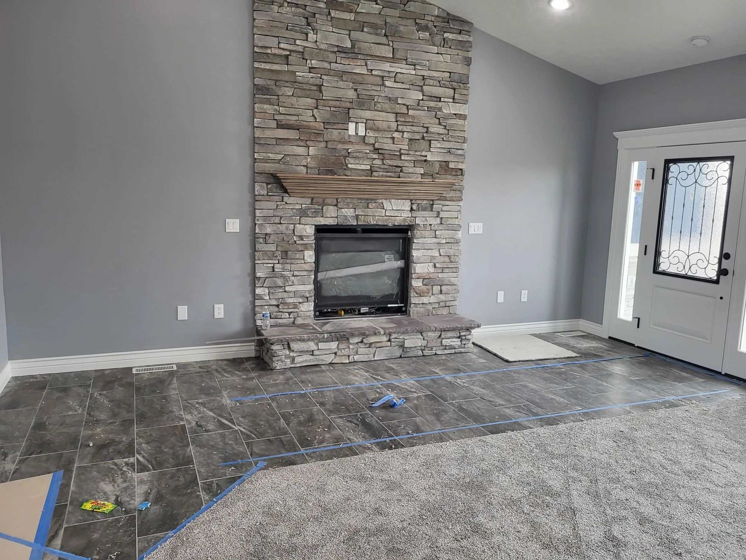 Living room with stone fireplace, gray walls, and a white front door with glass and decorative ironwork. The floor in front of the fireplace has dark tiles, with blue painter's tape marking the area. A water bottle and some small items are on the til