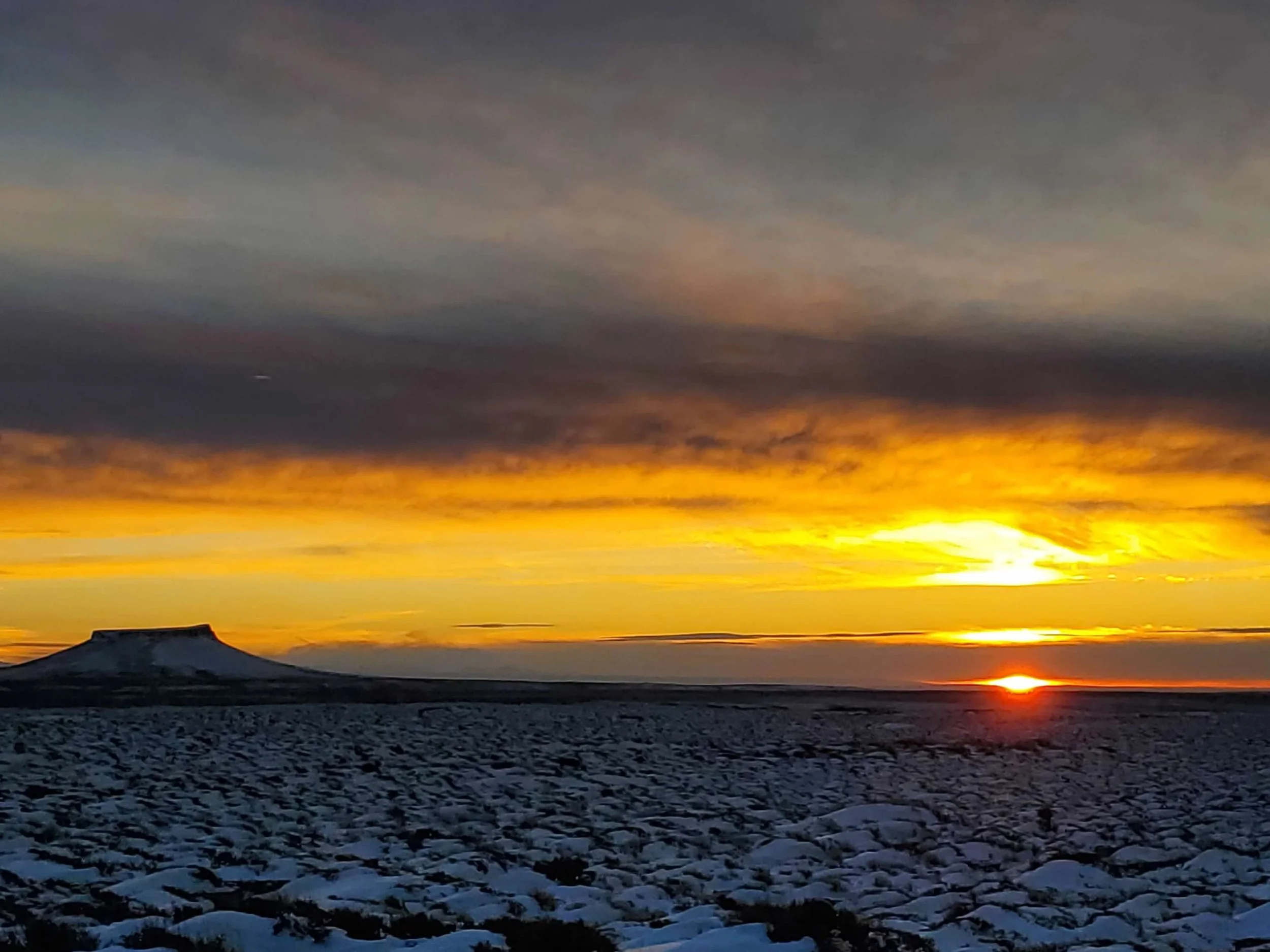 Sunset over a snow-covered landscape with a flat-topped mountain in the distance, orange and yellow hues in the sky.