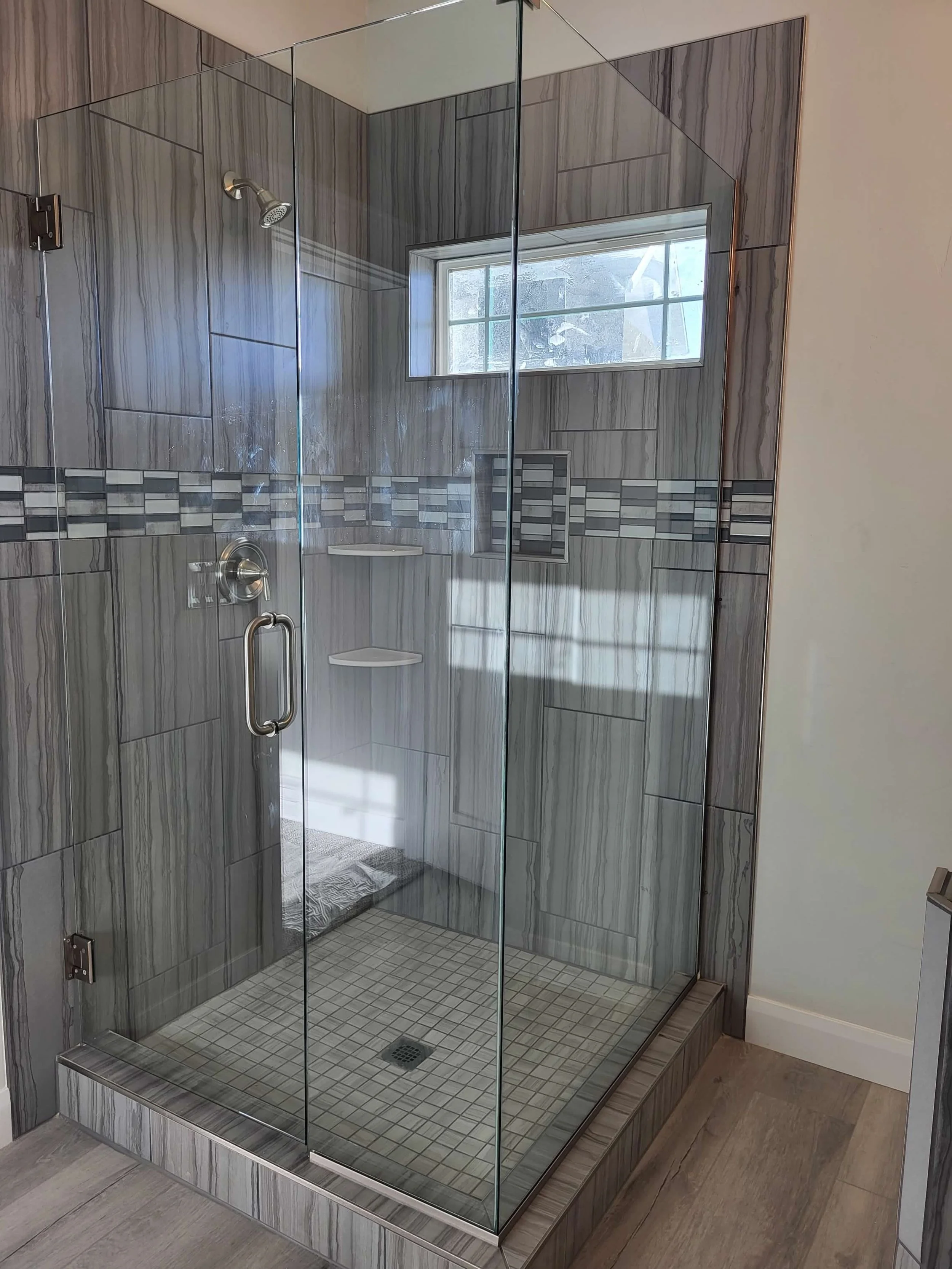 Glass shower enclosure with gray marble tiles, a horizontal decorative tile strip, a small window, a built-in soap shelf, and a floor drain.