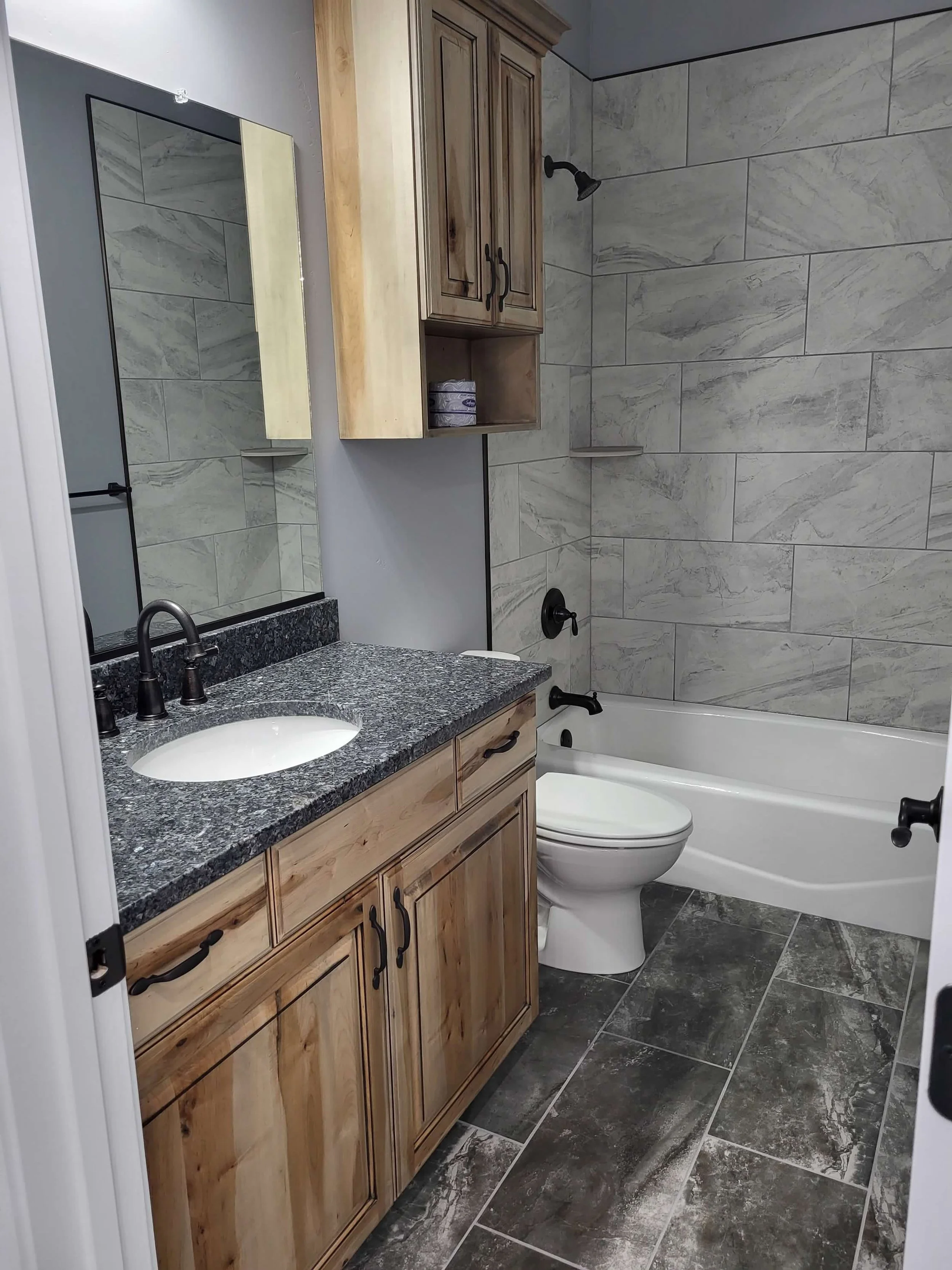 Bathroom with wooden cabinet vanity, granite countertop, mirror, bathtub with black fixtures, toilet, tiled wall and floor, and wall-mounted cabinet.
