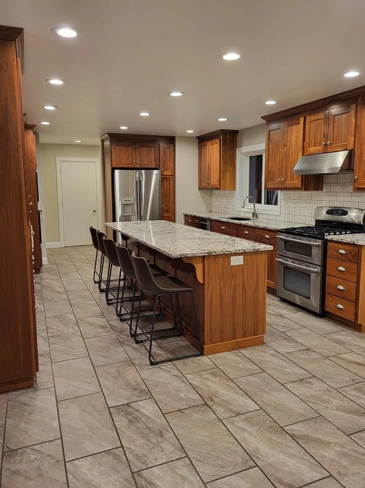 A spacious kitchen with wooden cabinets, granite countertops, stainless steel appliances, a large island with seating and a row of black chairs, tiled floor, white subway tile backsplash, and recessed ceiling lights.