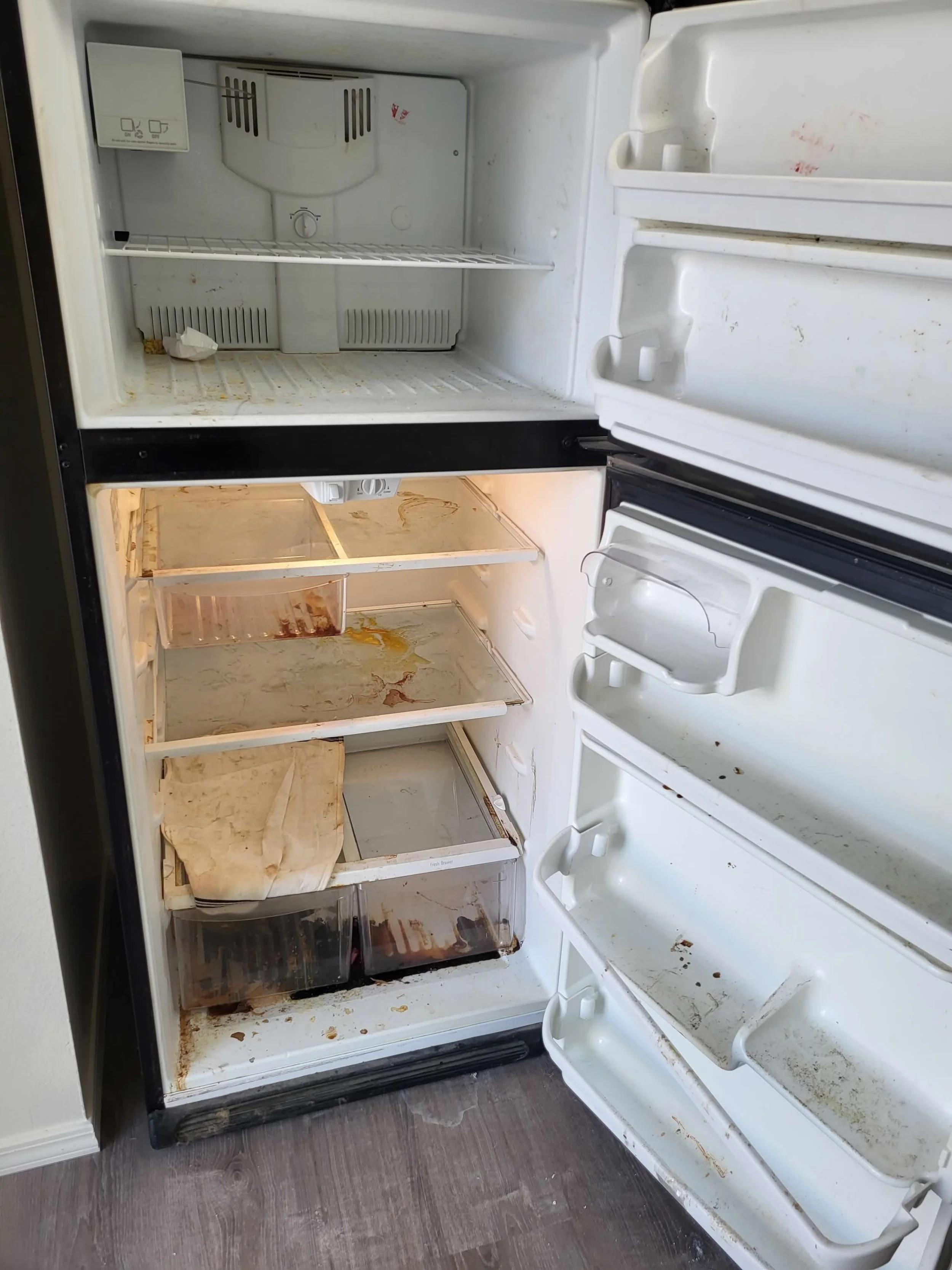 An open, dirty refrigerator with food stains, spilled liquids, and crumbs inside, including shelves and drawers.