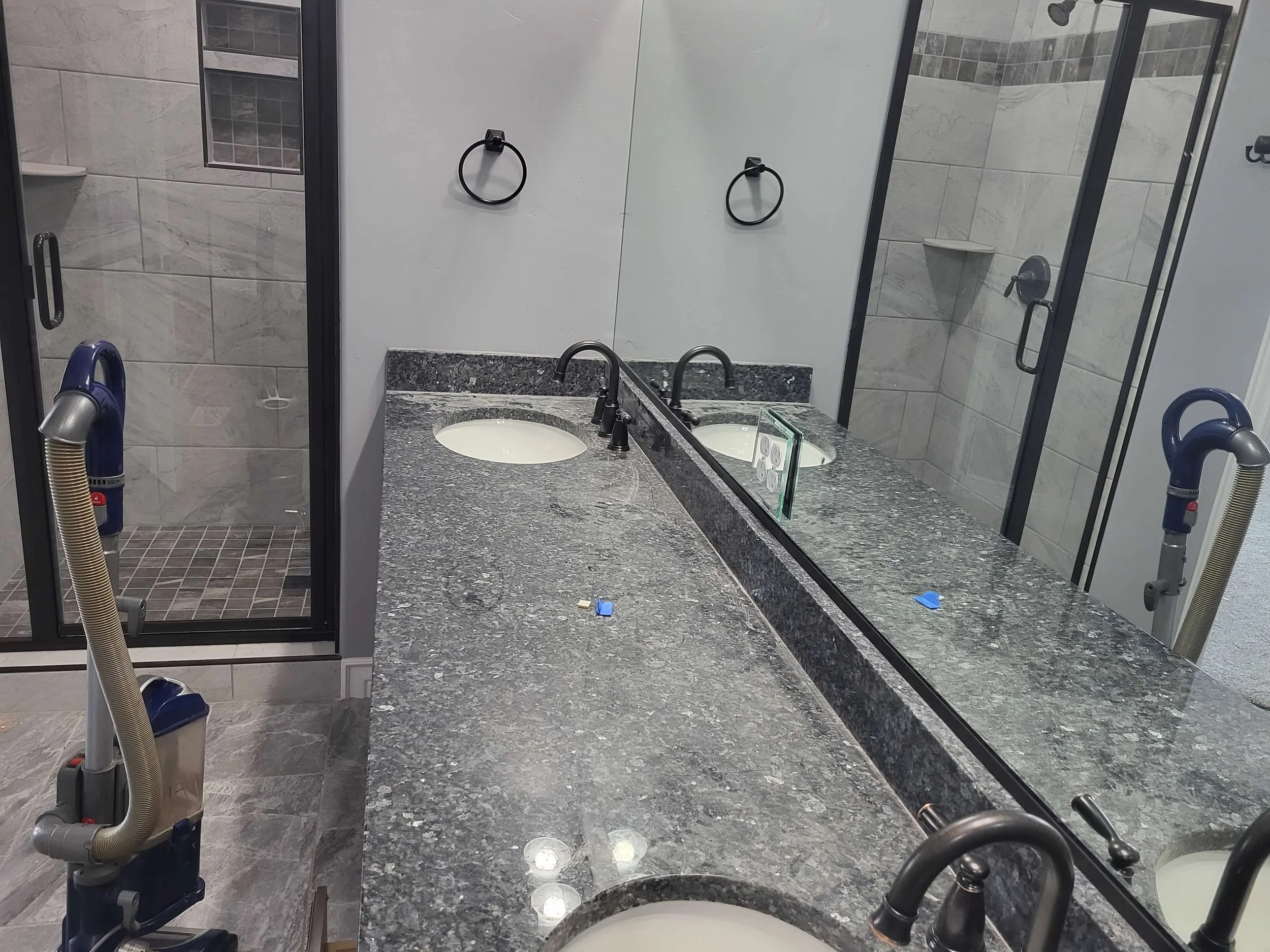 Empty bathroom with two sinks, granite countertop, large mirror, shower with glass door, and vacuum cleaner on the floor.