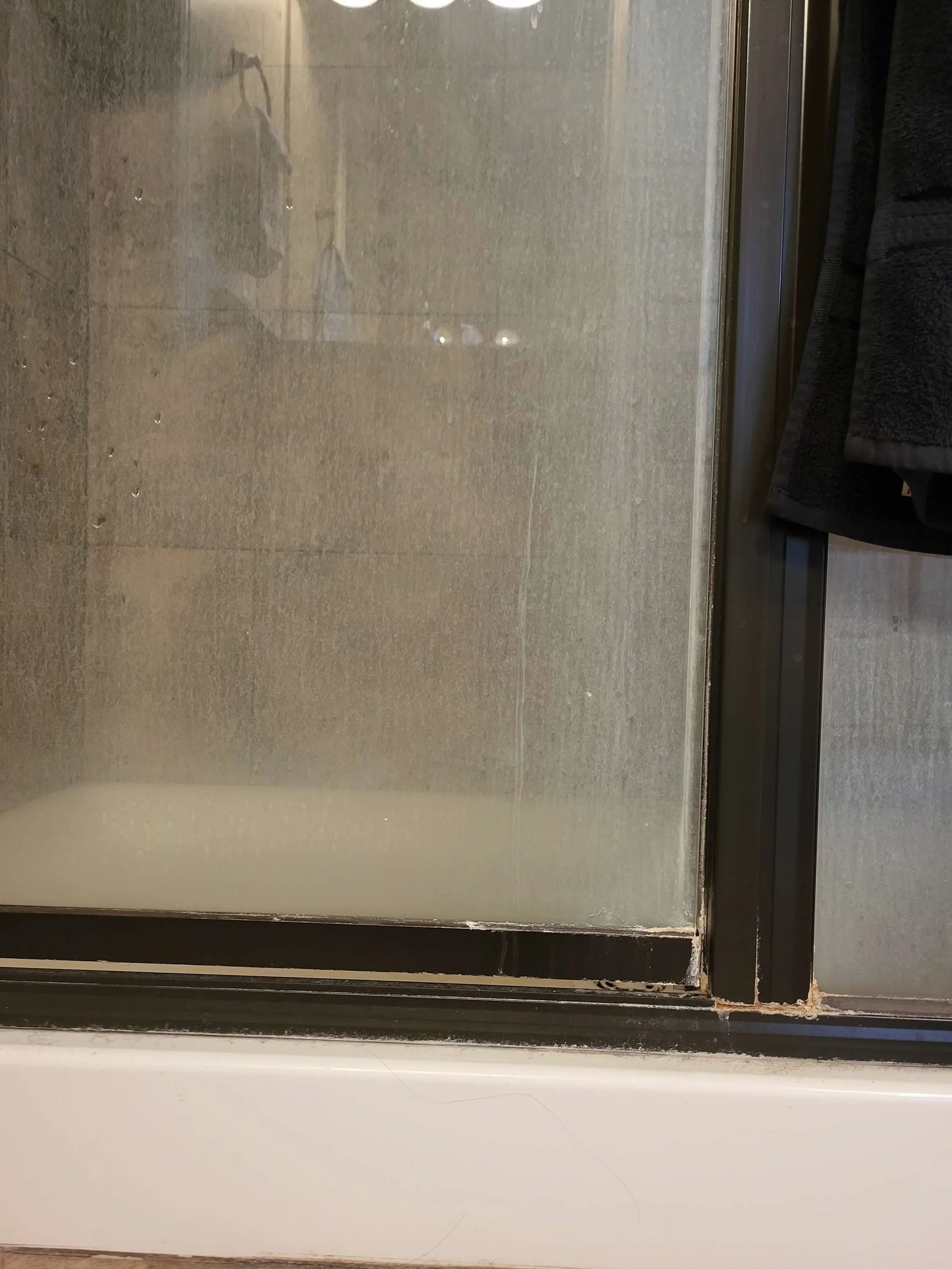 Close-up of a glass shower door with black metal trim and a beige tiled shower interior behind it. Part of a dark-colored towel hanging on a hook is visible on the right side.